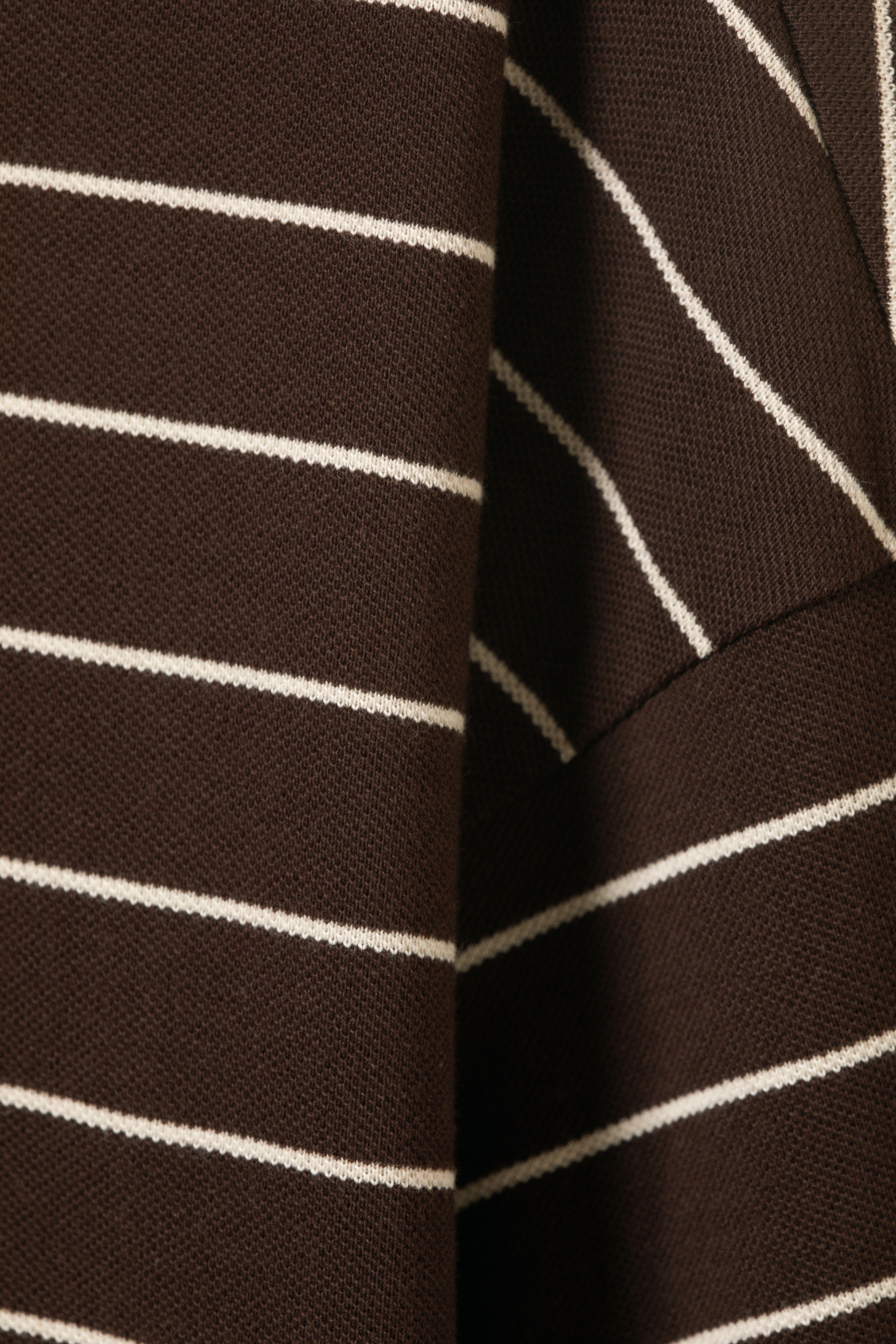 Größeres Bild ansehen: Ein langärmliges Poloshirt wird in einer Nahaufnahme gezeigt, das seinen dunkelbraunen Stoff mit dünnen, horizontalen weißen Streifen präsentiert. Der Stoff zeigt eine Piqué-Strickstruktur und ist gefaltet, wodurch sichtbare Tiefe entsteht.