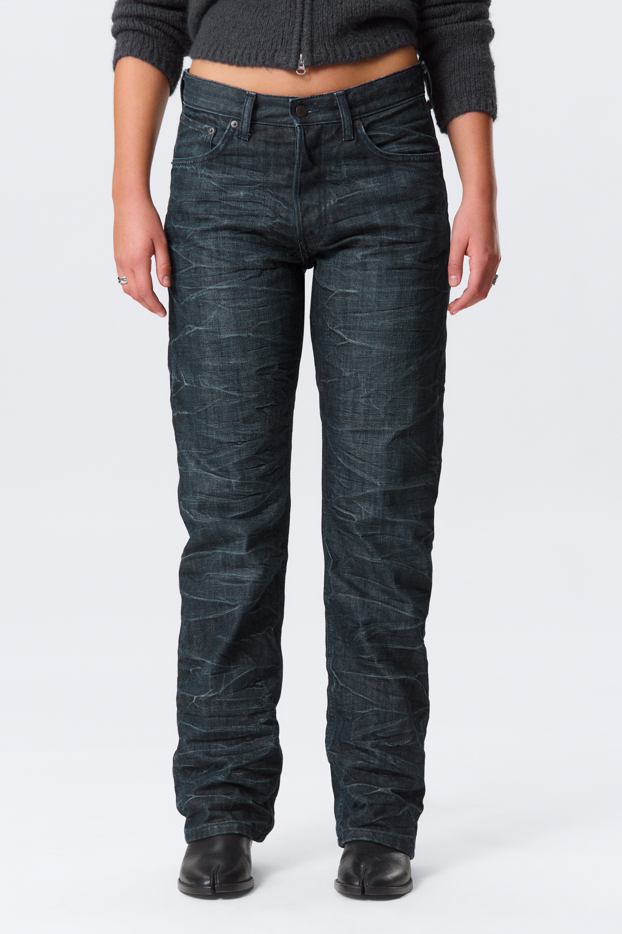 Se større billede: Mid-rise, lige ben jeans i mørkegrå denim bæres af en model. Jeansene har en falmet, krøllet struktur, en knap- og lynlåslukning samt synlige forlommer. Modellen har også en mørkegrå, kort sweater og sorte split-tå sko på.