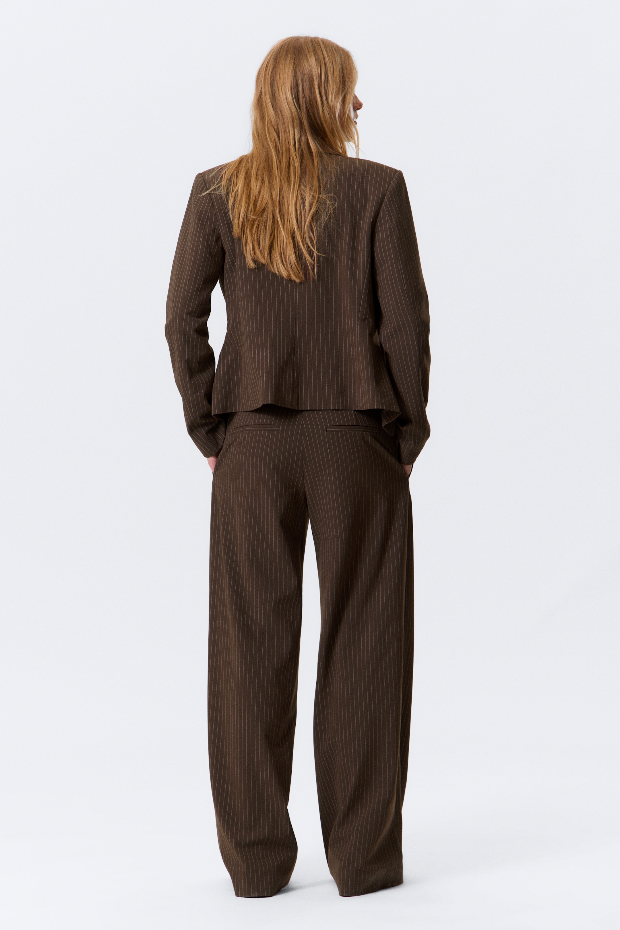 View larger image: A brown pinstriped suit is being presented from a back view, showing a cropped blazer and full-length, wide-leg trousers. The blazer features long sleeves, and the trousers are displaying two horizontal back pockets.