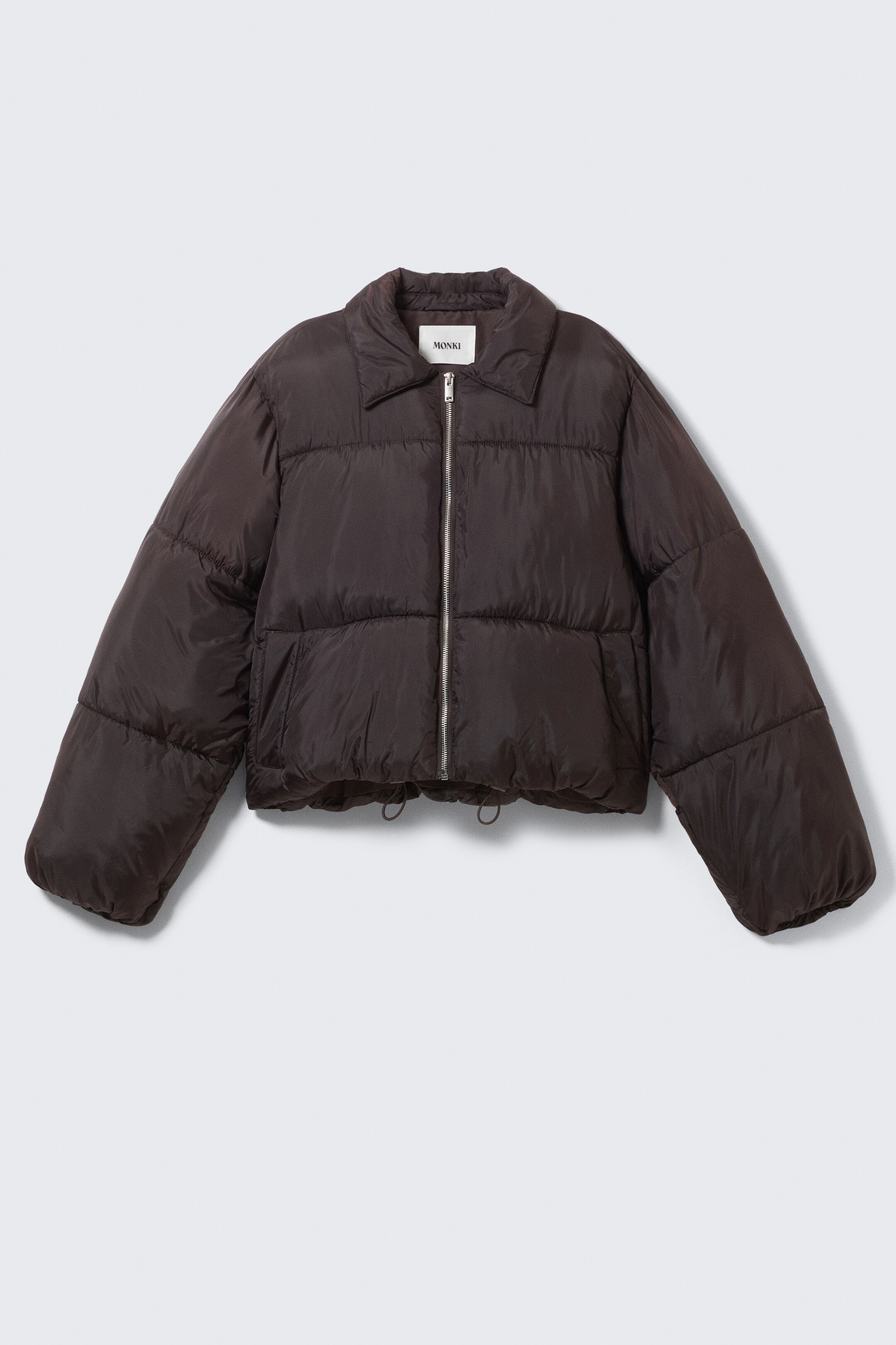View larger image: A dark brown cropped puffer jacket is being presented from the front, featuring a spread collar, a silver full-length zipper, and horizontal quilting across its body and long puffed sleeves. A drawstring is visible at the bottom hem, and a "MONKI" label is showing at the back of the neck.