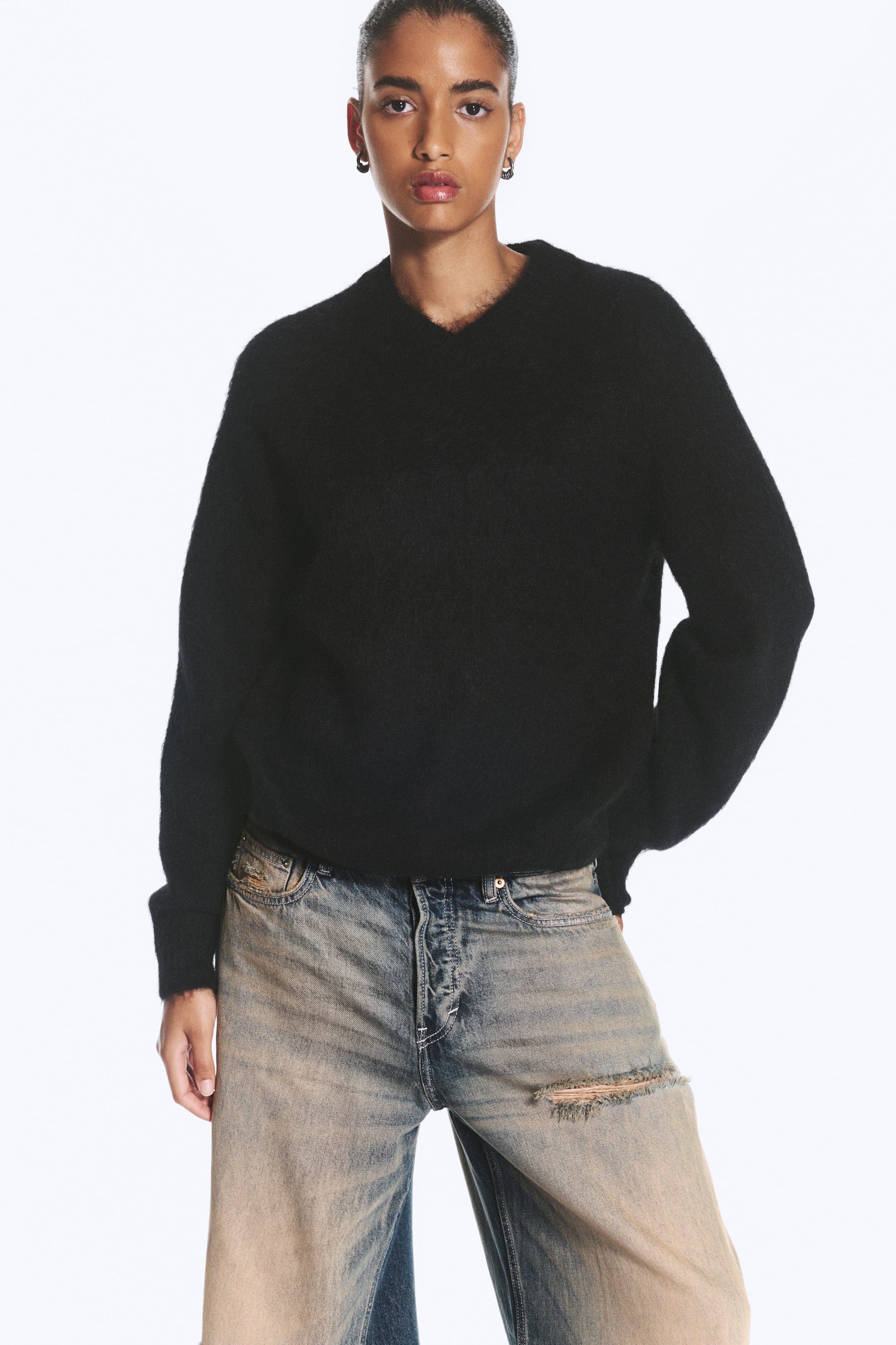 Grösseres Bild ansehen: Ein schwarzer, gestrickter Pullover aus Mohair-Mischung mit V-Ausschnitt, flauschiger Struktur und lockerer Passform wird vom Model getragen. Der Pullover ist mit einer hell gewaschenen Jeans im Used-Look kombiniert.