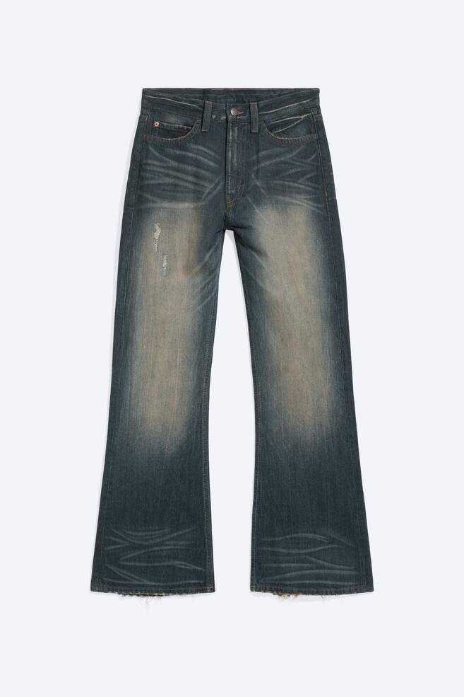 Een paar donkergewassen bootcut jeans wordt van voren getoond, met een lage taille, opvallende zandkleurige vervaging en whiskerdetails aan de heupen en zoom. De jeans hebben een kleine distressed scheur op de rechterbovenbeen en licht gerafelde onafgewerkte zomen. - 1