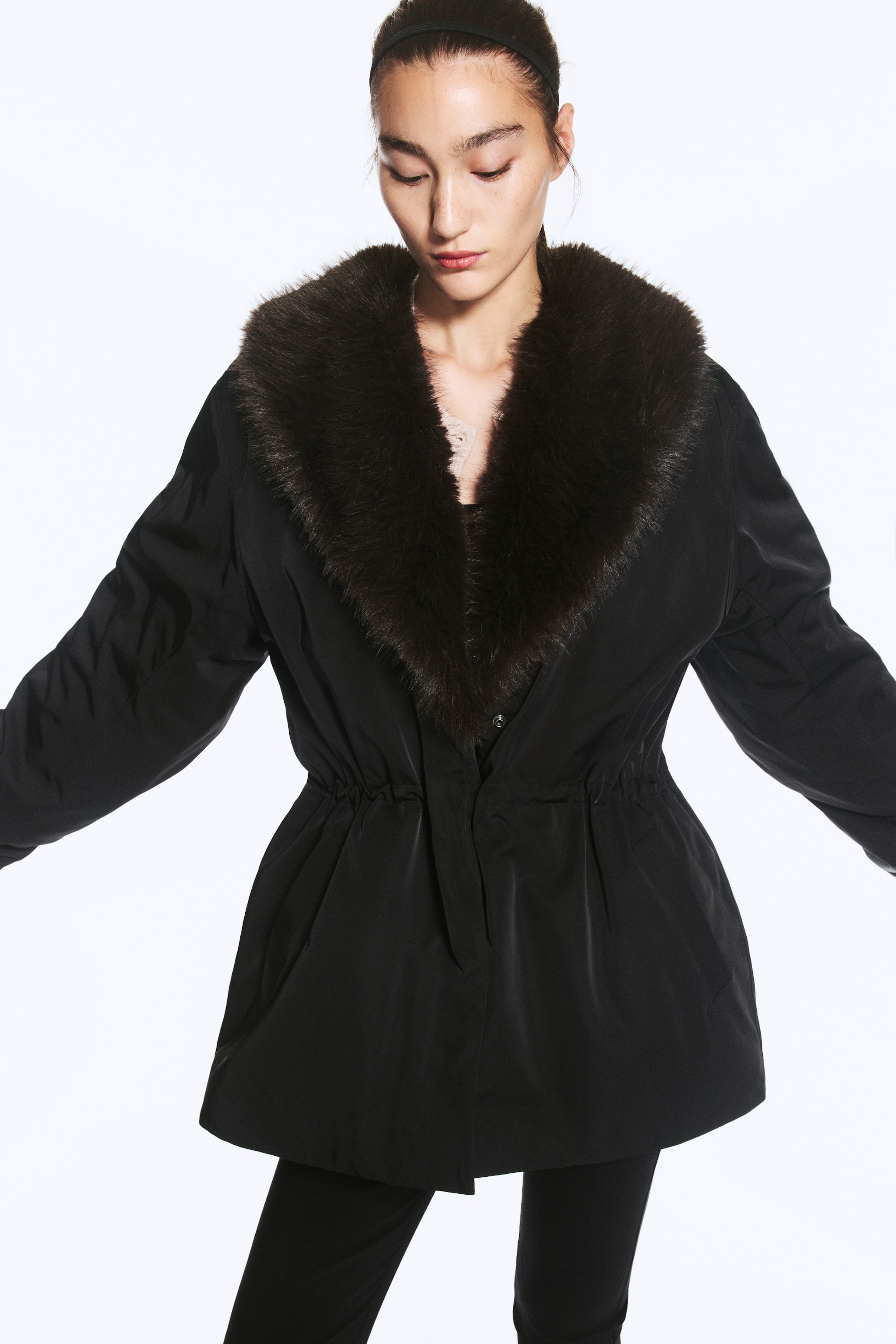 View larger image: A black padded jacket featuring a dark faux fur shawl collar and a cinched elasticated waist is being worn by the model. The jacket has a concealed front fastening and voluminous sleeves, styled with matching black trousers.
