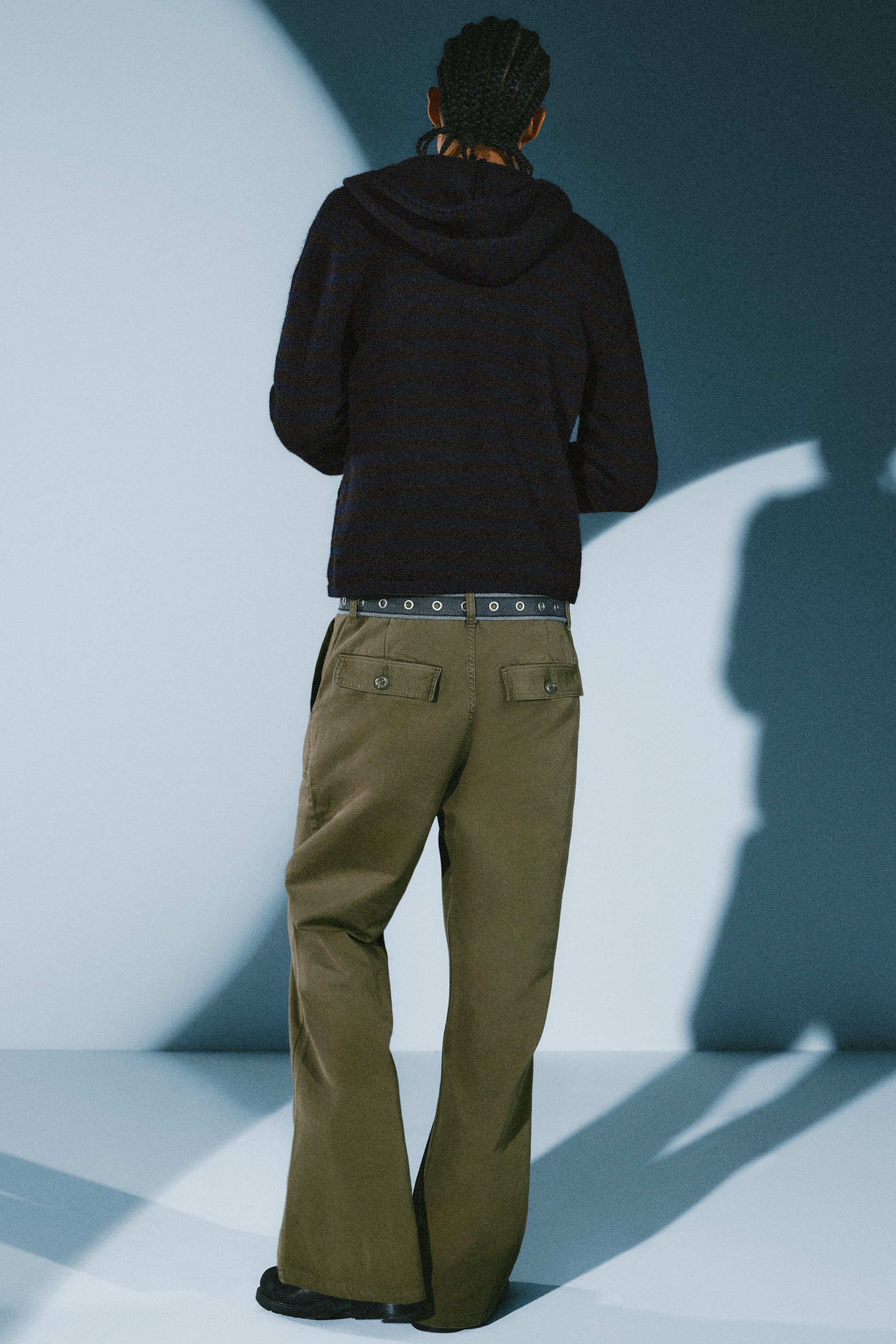 Größeres Bild anzeigen: Eine olivgrüne Arbeitshose mit Bootcut-Bein wird vom Model getragen und setzt die hinten mit Patten und Knöpfen versehenen Taschen in Szene. Die Low-Rise-Hose ist mit einem grauen Gürtel mit Ösen und einem dunklen Kapuzenpullover kombiniert.