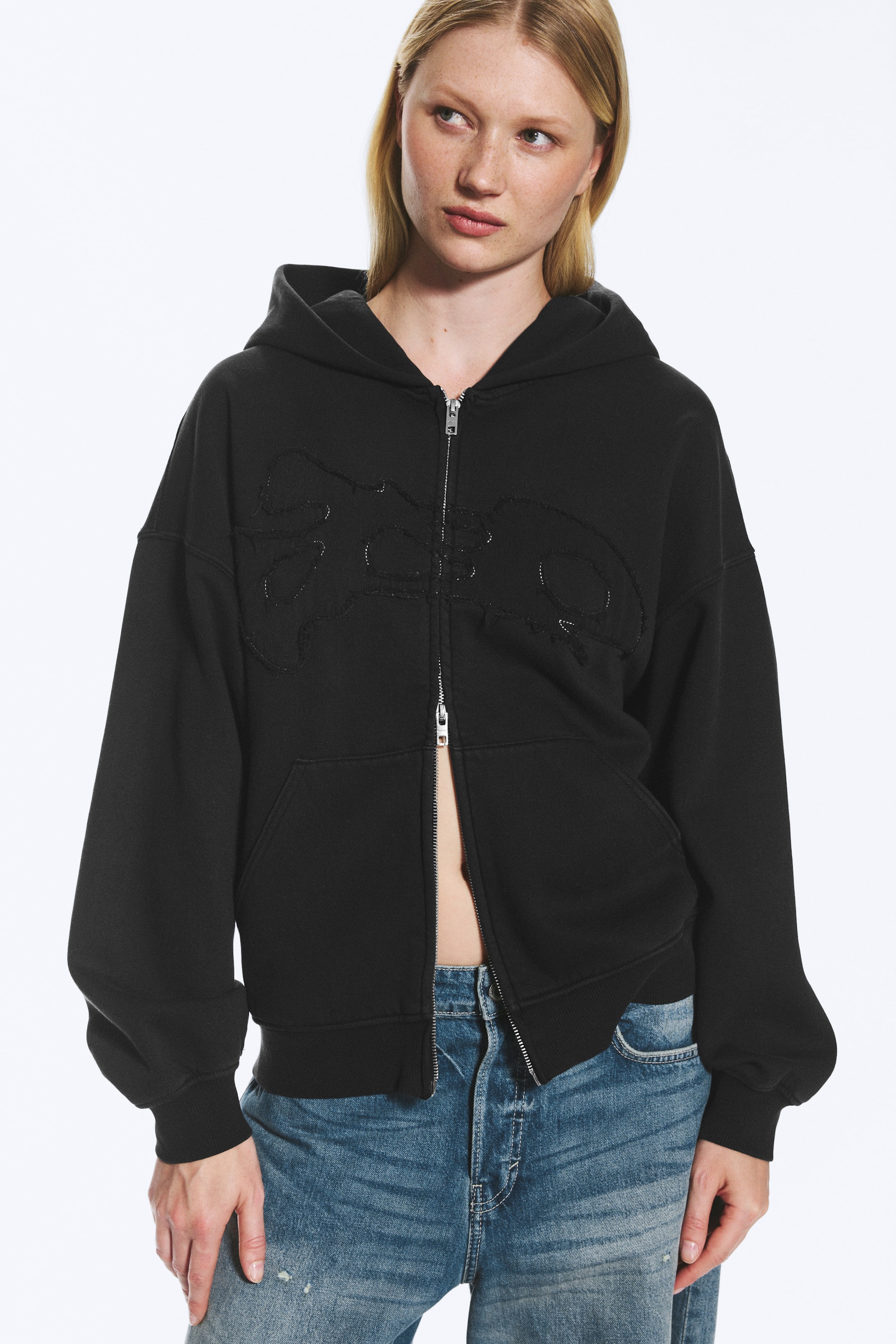 View larger image: The model is wearing an oversized black zip-up hoodie, featuring dropped shoulders, long sleeves with ribbed cuffs, a ribbed hem, and two front pockets. A subtle stitched graphic detail is visible on the chest, and the zip is partially open, styled with blue jeans.
