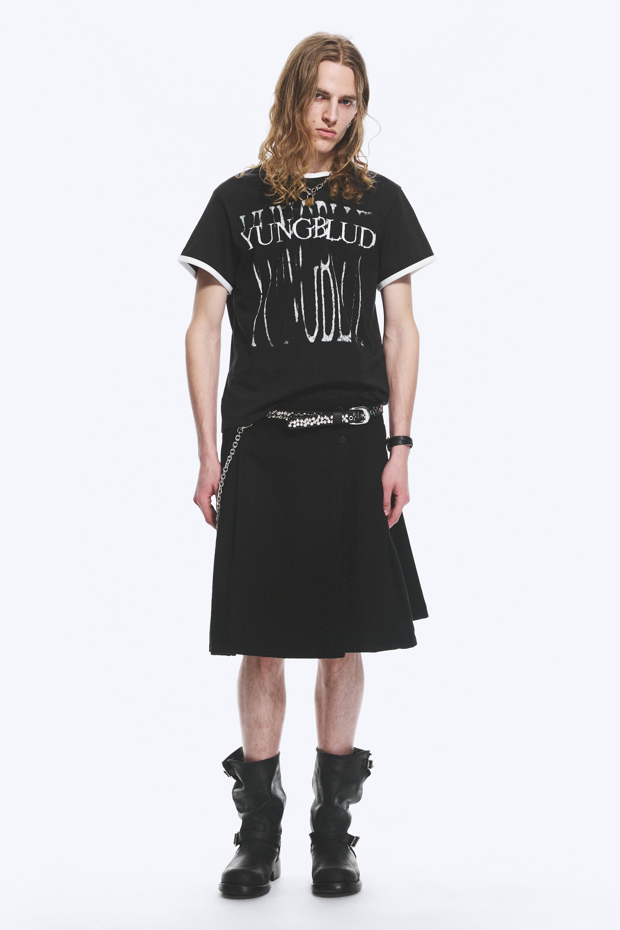 Nagyobb kép megtekintése: Egy fekete, rövid ujjú póló, amelyen egy kopott fehér 'YUNGBLUD' grafika látható, fehér szegéllyel a galléron és az ujjak mandzsettáján, egy fekete térdig érő kilt-tel van viselve. A kiltet egy szegecses öv díszíti, amelyről egy ezüst lánc lóg, és fekete, közép-vádliig érő csizmák csattos részletekkel egészítik ki a megjelenést.