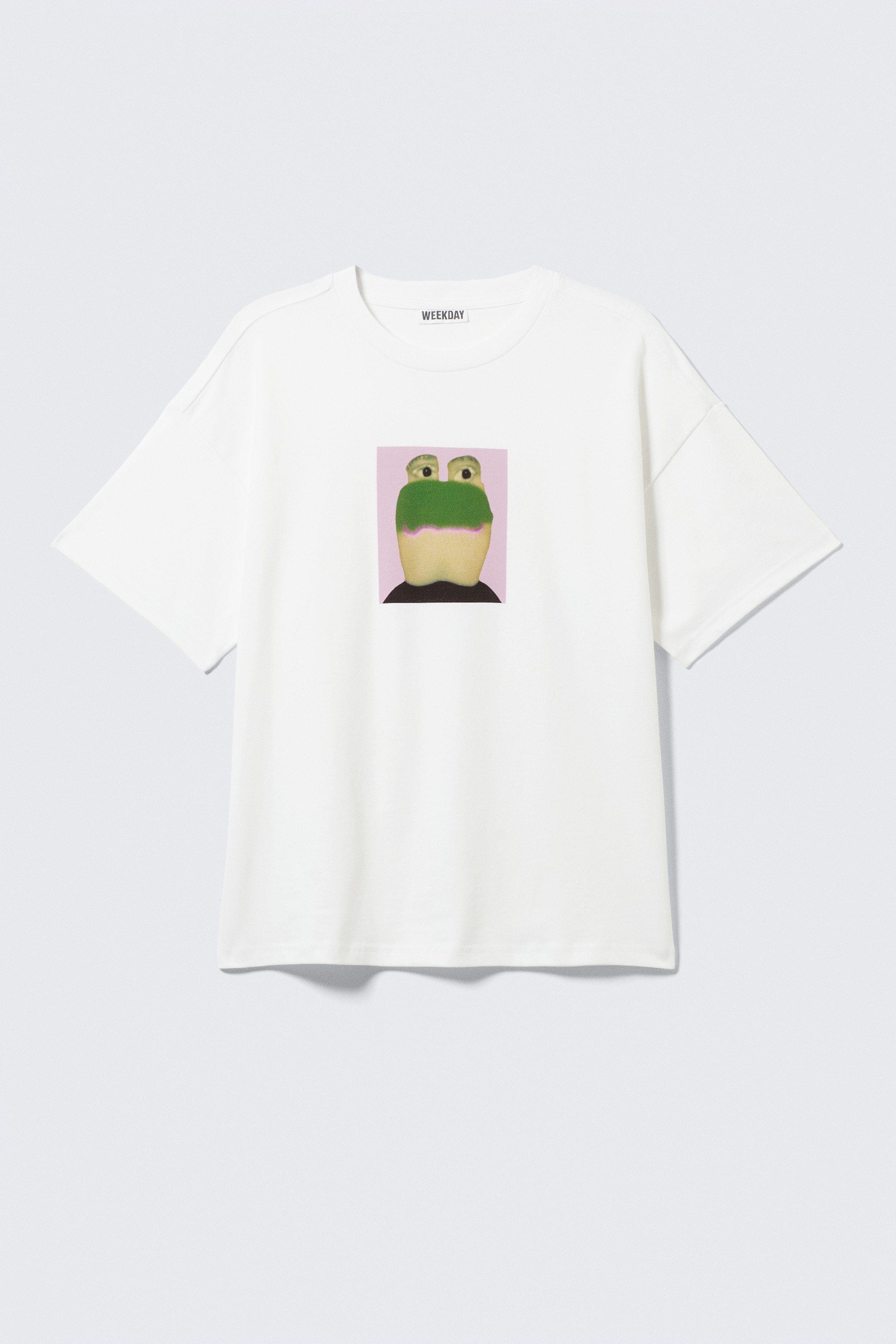 Agrandir l'image: Un t-shirt blanc affiche une coupe ample et carrée avec des épaules tombantes et un col arrondi, où une étiquette 'WEEKDAY' est visible. Sur son DEVANT, un graphisme carré montre le visage stylisé d'une créature avec de grands yeux, une tête supérieure verte et un visage inférieur de couleur plus claire, sur un fond violet clair.