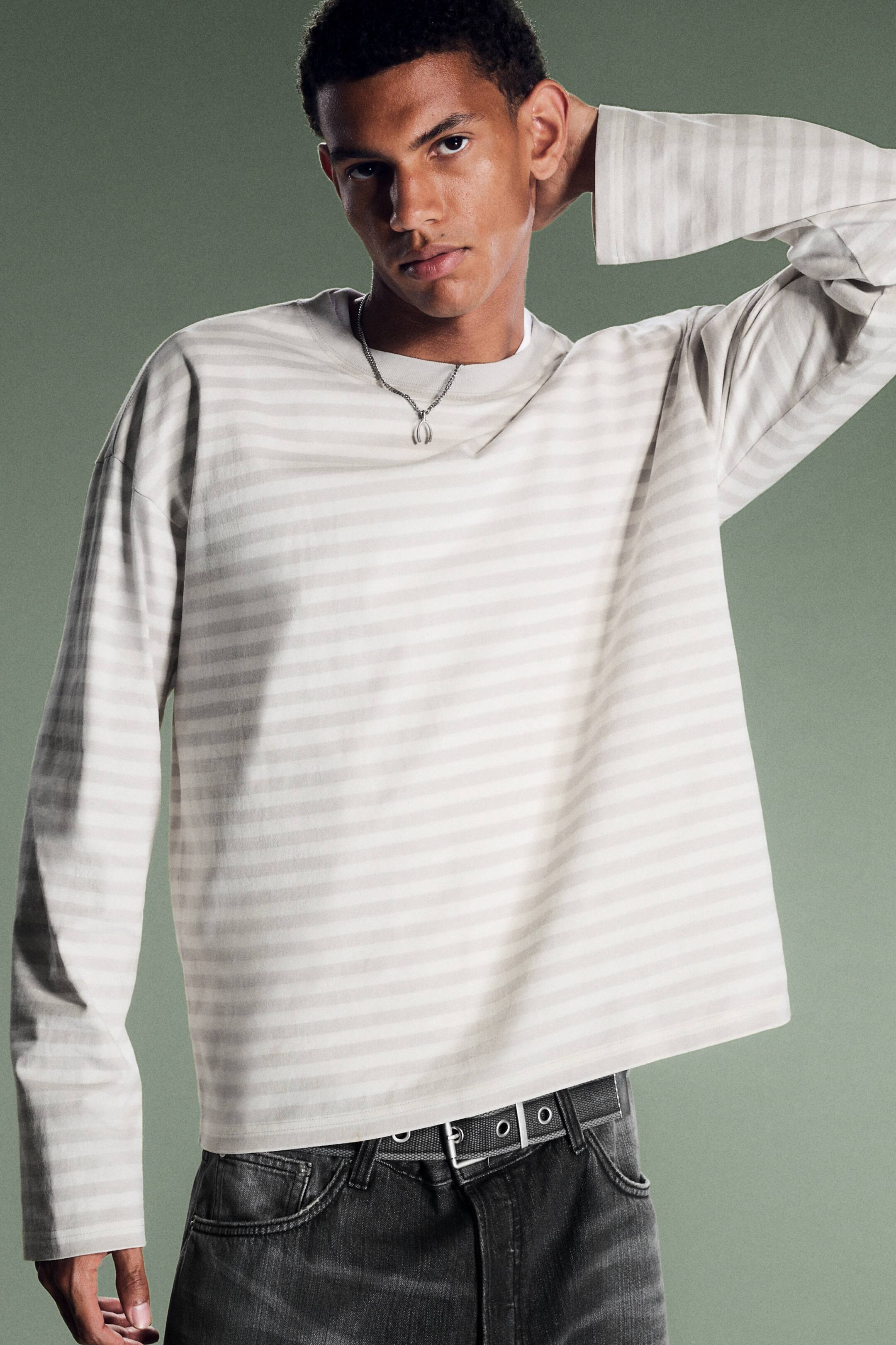 Größeres Bild ansehen: Ein kastig geschnittenes T-Shirt mit langen Ärmeln und dezenten horizontalen Streifen wird vom Model getragen. Das locker sitzende Shirt hat einen Rundhalsausschnitt und einen geraden Saum und ist mit dunkelgrauen Jeans und einer silbernen Halskette kombiniert.