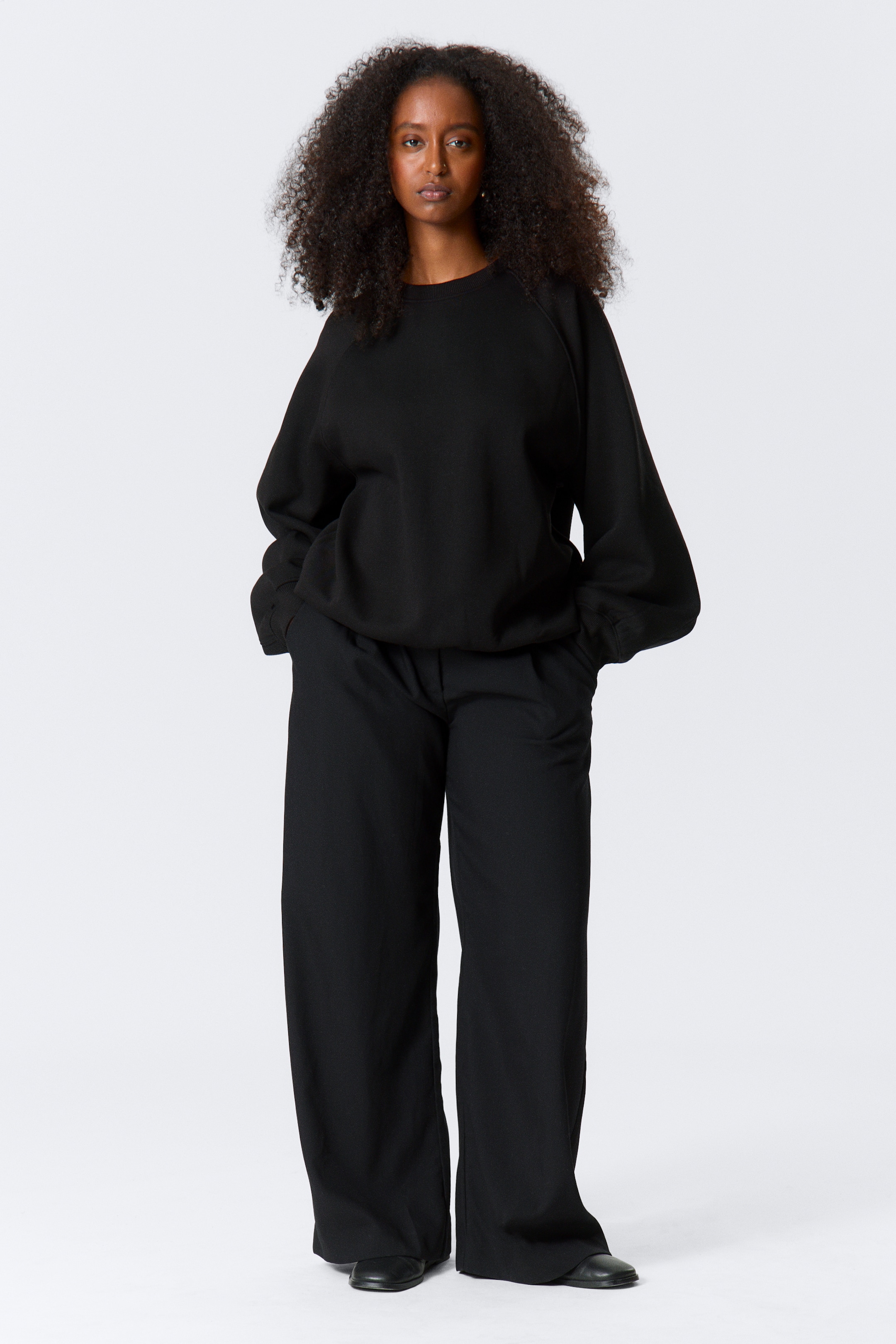 Größeres Bild anzeigen: Ein schwarzes, übergroßes Crewneck-Sweatshirt mit Raglanärmeln, gerippten Bündchen und einem gerippten Saum wird frontseitig getragen. Es wird mit schwarzen, weit geschnittenen Hosen kombiniert, die vollständig bis zum Boden fallen und sichtbare Vordertaschen zeigen.