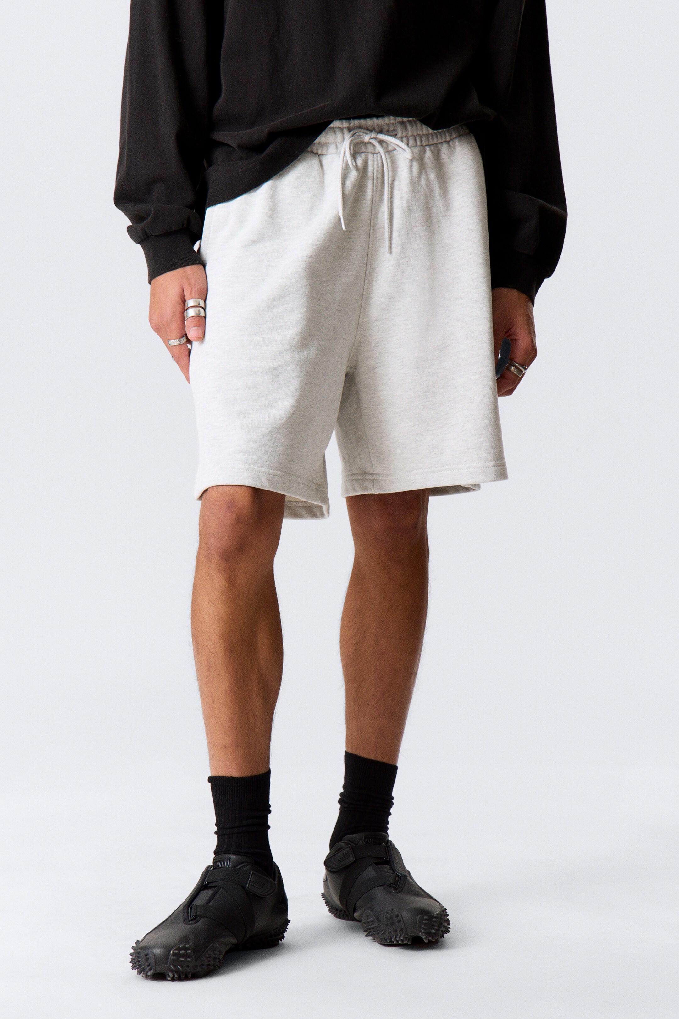 Grotere afbeelding bekijken: Lichtgrijze shorts worden gedragen, met een elastische tailleband en een witte trekkoord aan de voorkant. De shorts hebben een ontspannen pasvorm, reiken tot boven de knie en worden aan de bovenkant gedeeltelijk bedekt door een zwart shirt met lange mouwen.