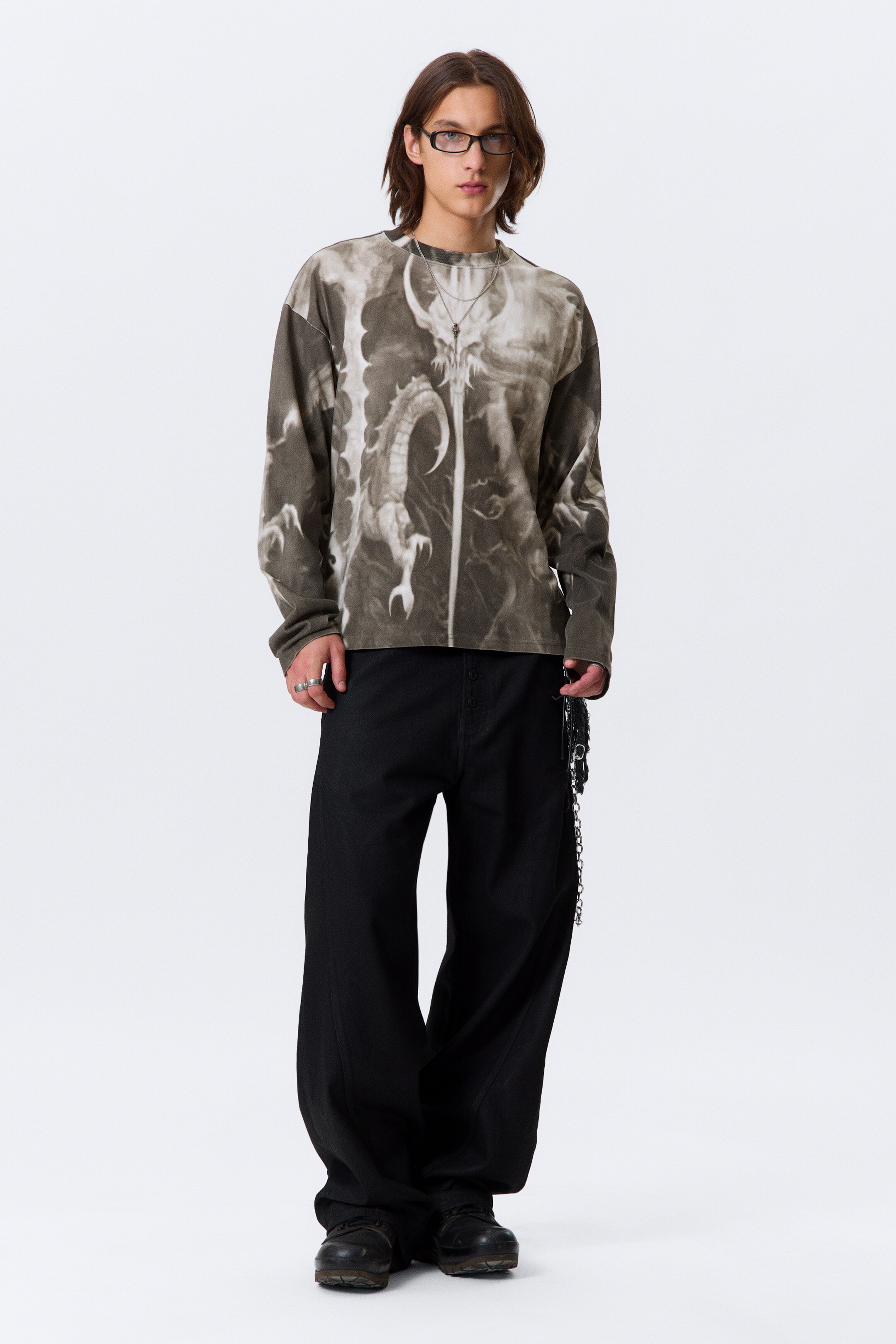 Agrandir l'image: Un T-shirt à manches longues et à coupe carrée, fabriqué en jersey de coton, est présenté de face, arborant un grand imprimé graphique estompé d'une créature semblable à un dragon dans des tons atténués sur la poitrine et les manches. Le T-shirt est conçu avec un col rond côtelé et des ourlets à double couture.