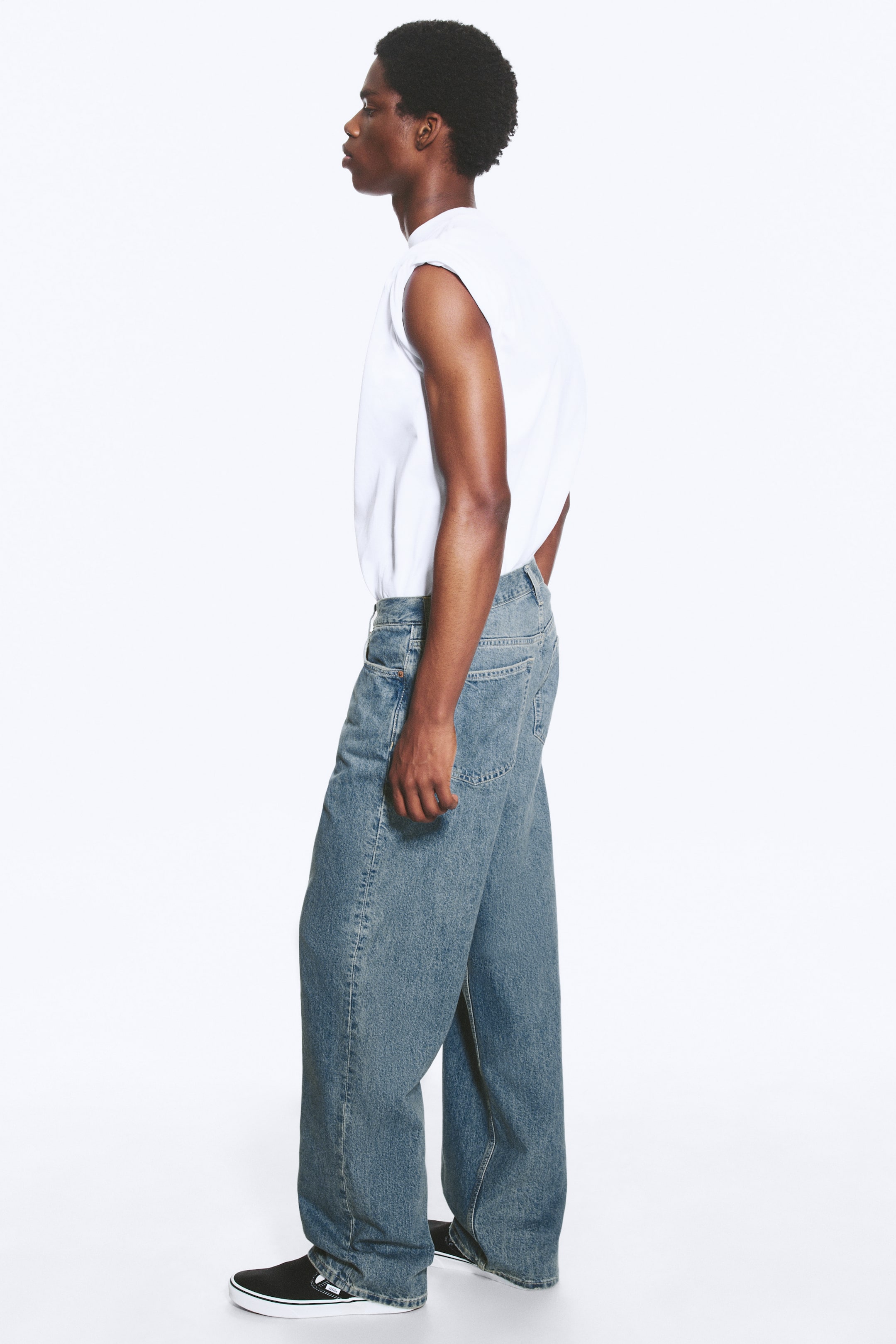 Vis større bilde: Lyseblå, mid-rise jeans med en løs, rett benpassform bæres av modellen. Jeansene har et klassisk fem-lommers design. En hvit ermeløs topp er stukket inn i jeansens linning, og de brede benåpningene dekker svarte slip-on sko.