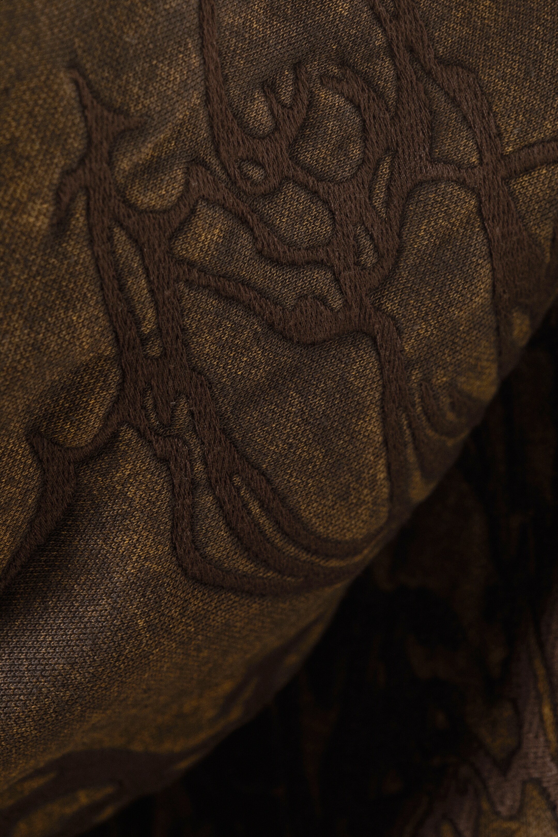 View larger image: An embroidered hoodie's fabric is displaying a raised, intricate pattern in dark brown thread. The swirling, organic design is contrasting with the brownish-olive knit material.