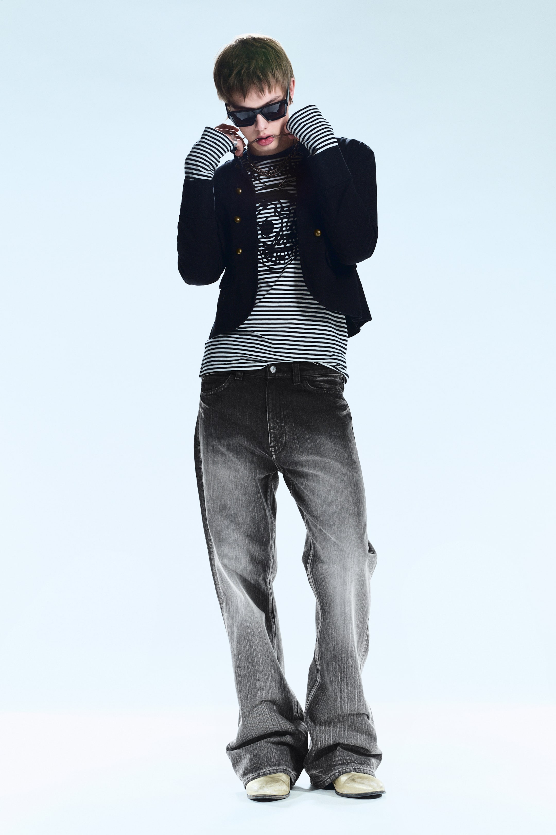 Zobacz większy obraz: Czarna kurtka do talii, zaprezentowana z przodu z dwoma rzędami guzików w złotym odcieniu, noszona jest otwarta na czarno-białej koszuli w paski z długimi rękawami, która prezentuje grafikę na klatce piersiowej, a jej mankiety w paski wystają z rękawów kurtki. Spodnie jeansowe o luźnym kroju, w wyblakłym szarym kolorze, z szerokimi nogawkami, opadają na jasne buty.