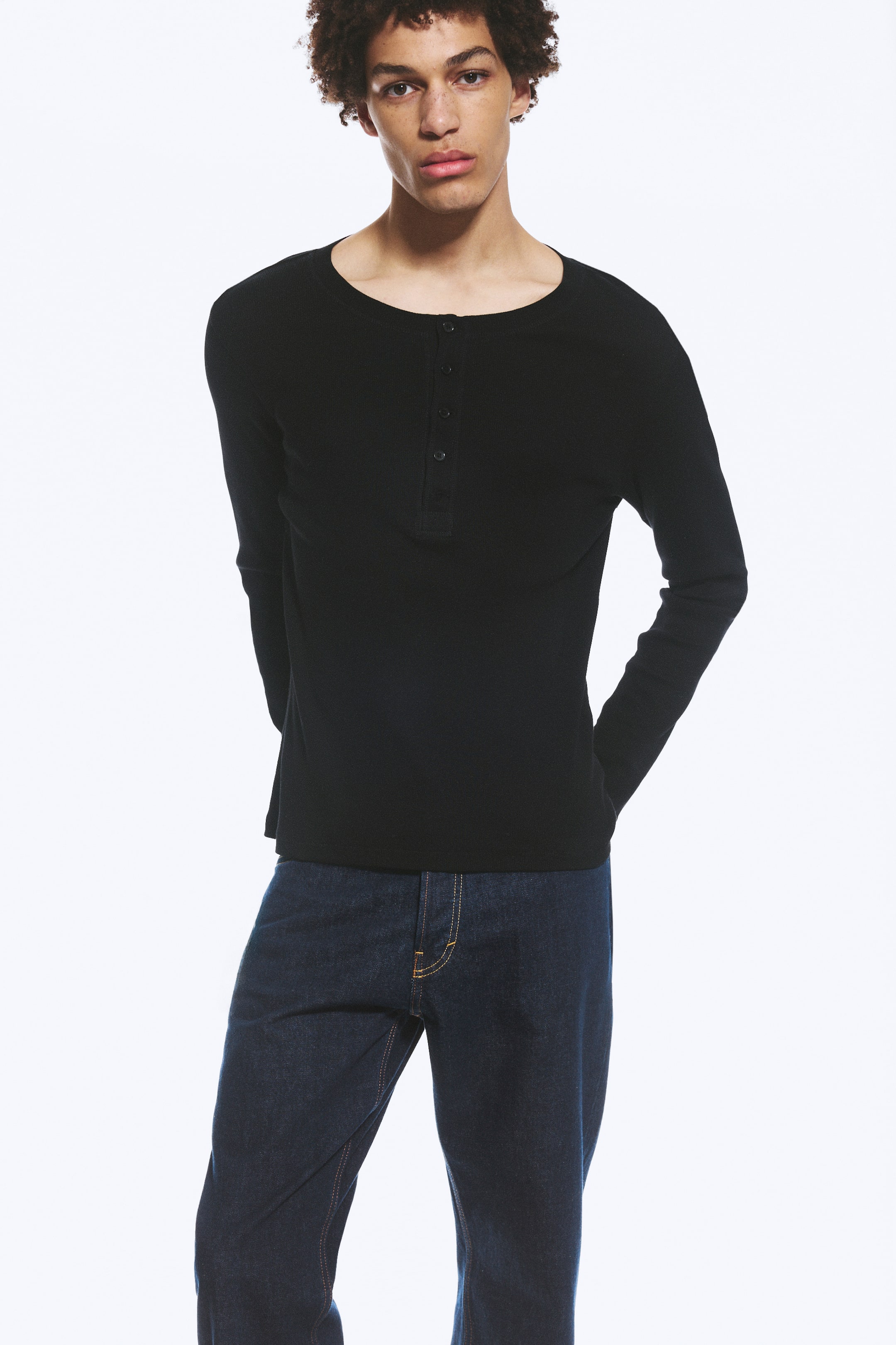 View larger image: A black long-sleeve Henley shirt is showcased, featuring a ribbed texture and a four-button placket at the neckline. It is paired with dark wash jeans, visible from the waist down, which display contrasting orange stitching.