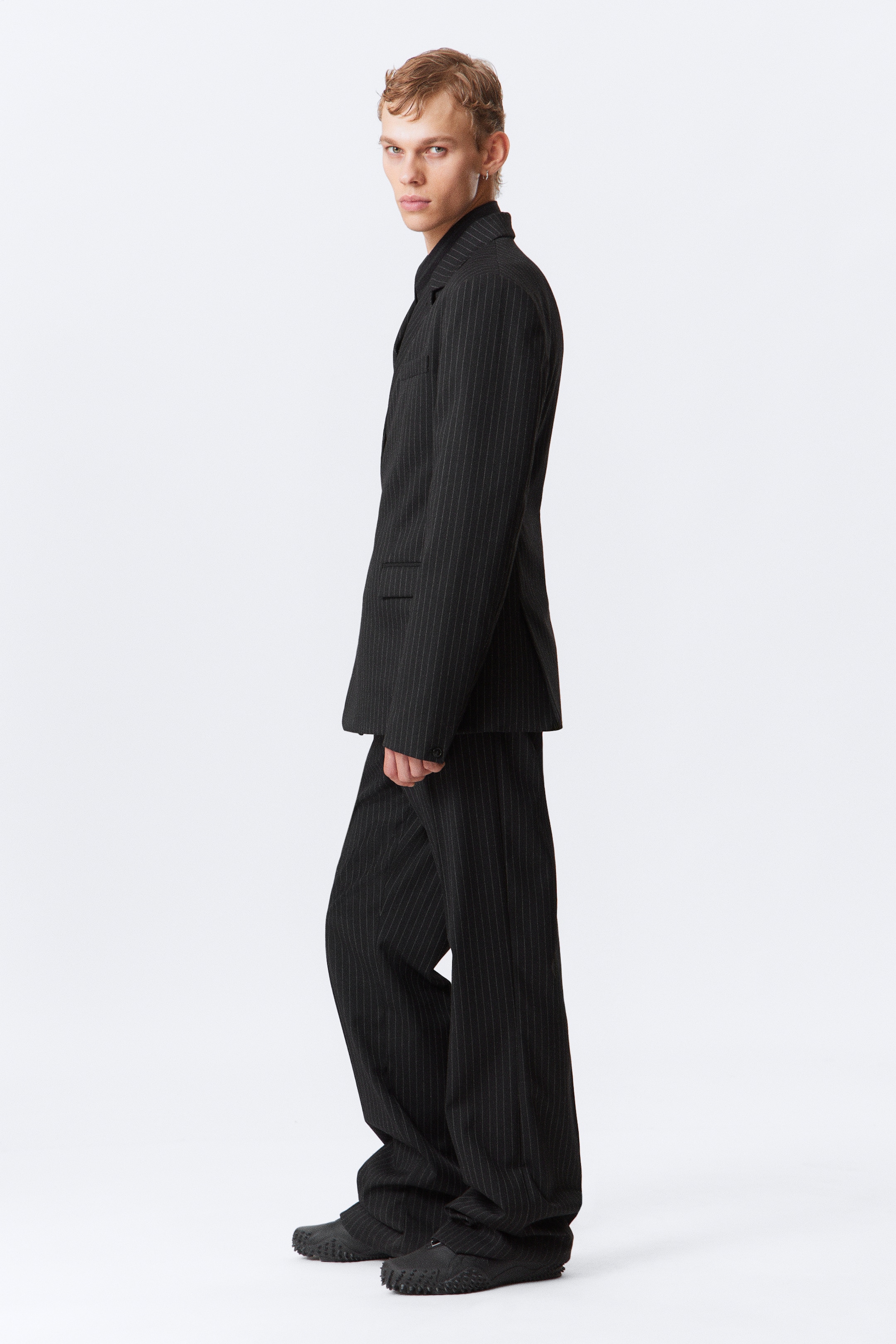 Agrandir l'image: Un costume à rayures fines foncées est présenté de profil gauche. La veste présente des revers en pointe et une poche à rabat, tandis que le pantalon coordonné affiche une coupe large et ample, tombant sur des chaussures noires texturées. Une chemise foncée est portée sous la veste.