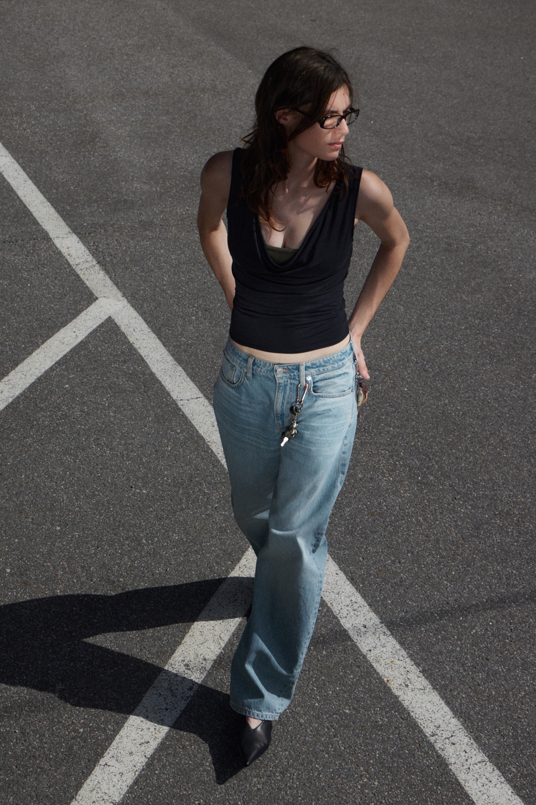 Se større billede: En person har en sort ærmeløs top med en draperet halsudskæring, lagdelt over et grønt undertøj. De har også lyse, vidde denimjeans på, hvorfra et sæt nøgler hænger fra en bæltestrop, og sorte spidse sko. Mørkeindfattede briller hviler på deres ansigt.