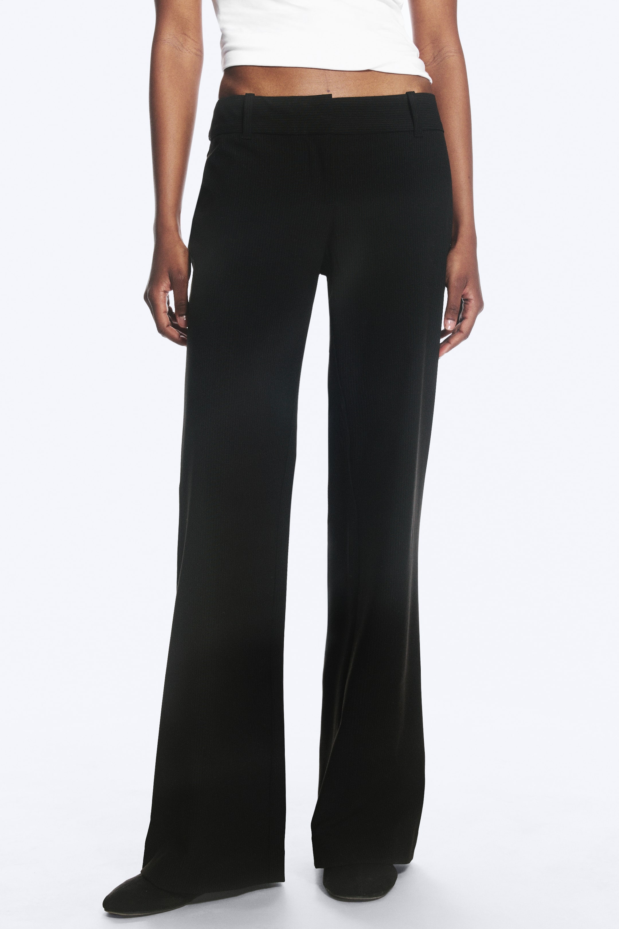 Agrandir l'image: Le mannequin porte un pantalon de costume noir sans poches, avec une texture côtelée, une taille basse avec passants de ceinture, une coupe ajustée au niveau des cuisses et une jambe légèrement évasée, assorti d'un haut court blanc.