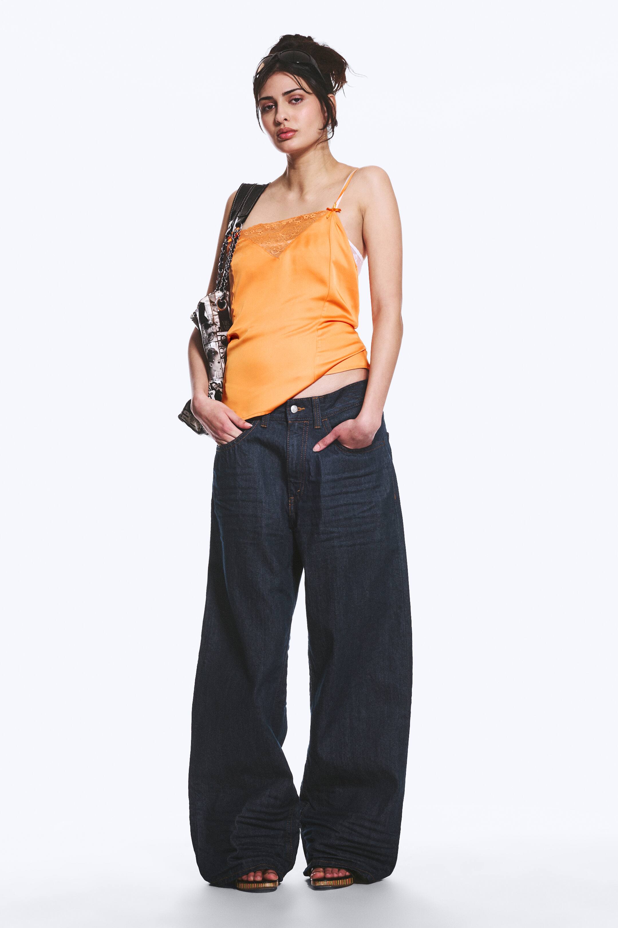 Se større billede: En orange satin camisole med en blondekantet V-udskæring og tynde spaghetti-stropper, med små sløjfer der pryder stropfæstningerne. Den er delvist stukket ned i mørkvaskede, brede jeans, som har en løs pasform og samler sig ved anklerne.