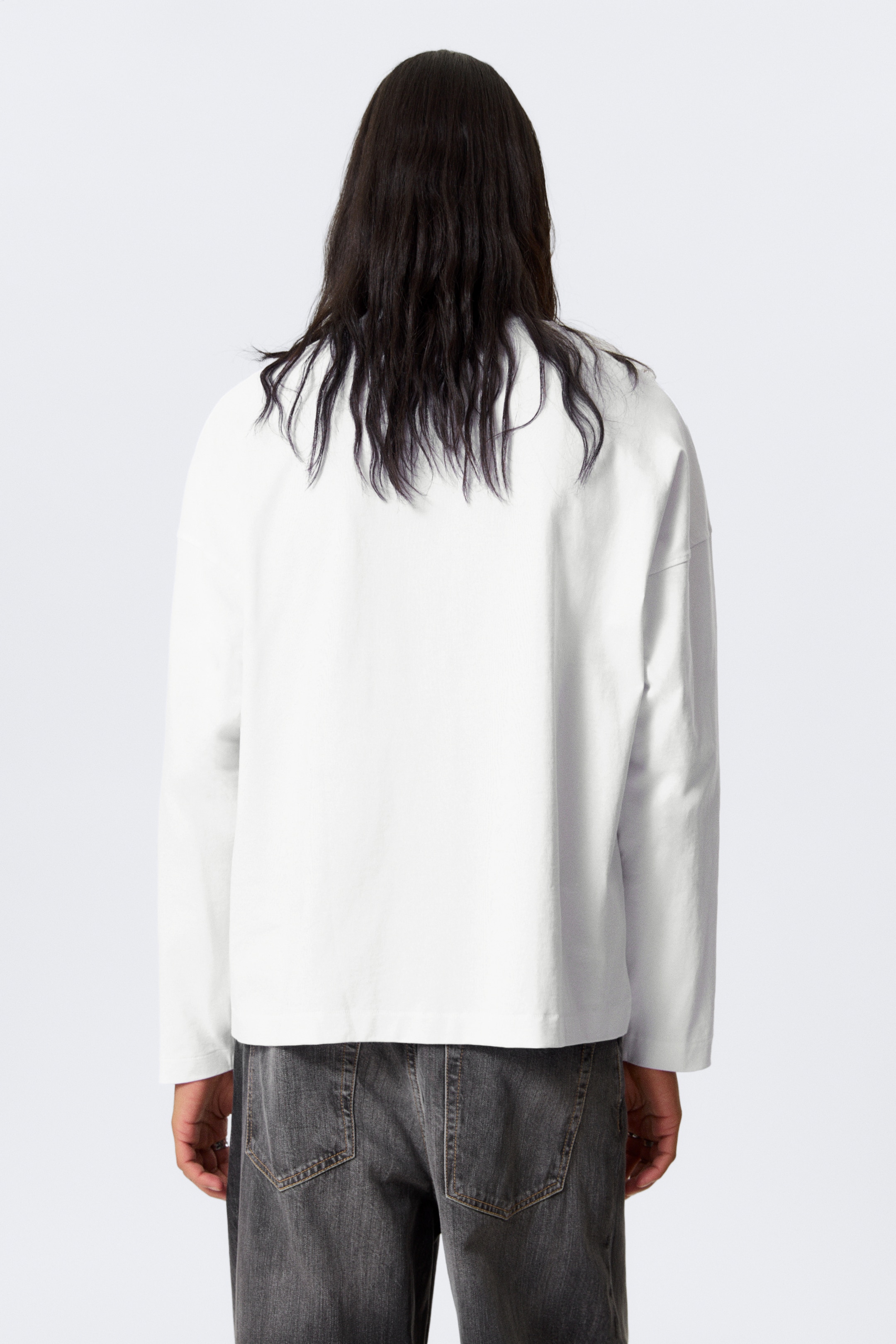 Visa större bild: En vit långärmad T-shirt ses från BAKSIDAN, och visar upp en överdimensionerad, boxig silhuett med en fyrkantig form och nedfallna axlar. Den har breda, avsmalnande ärmar, en rund halsringning och synliga sydda fållar.