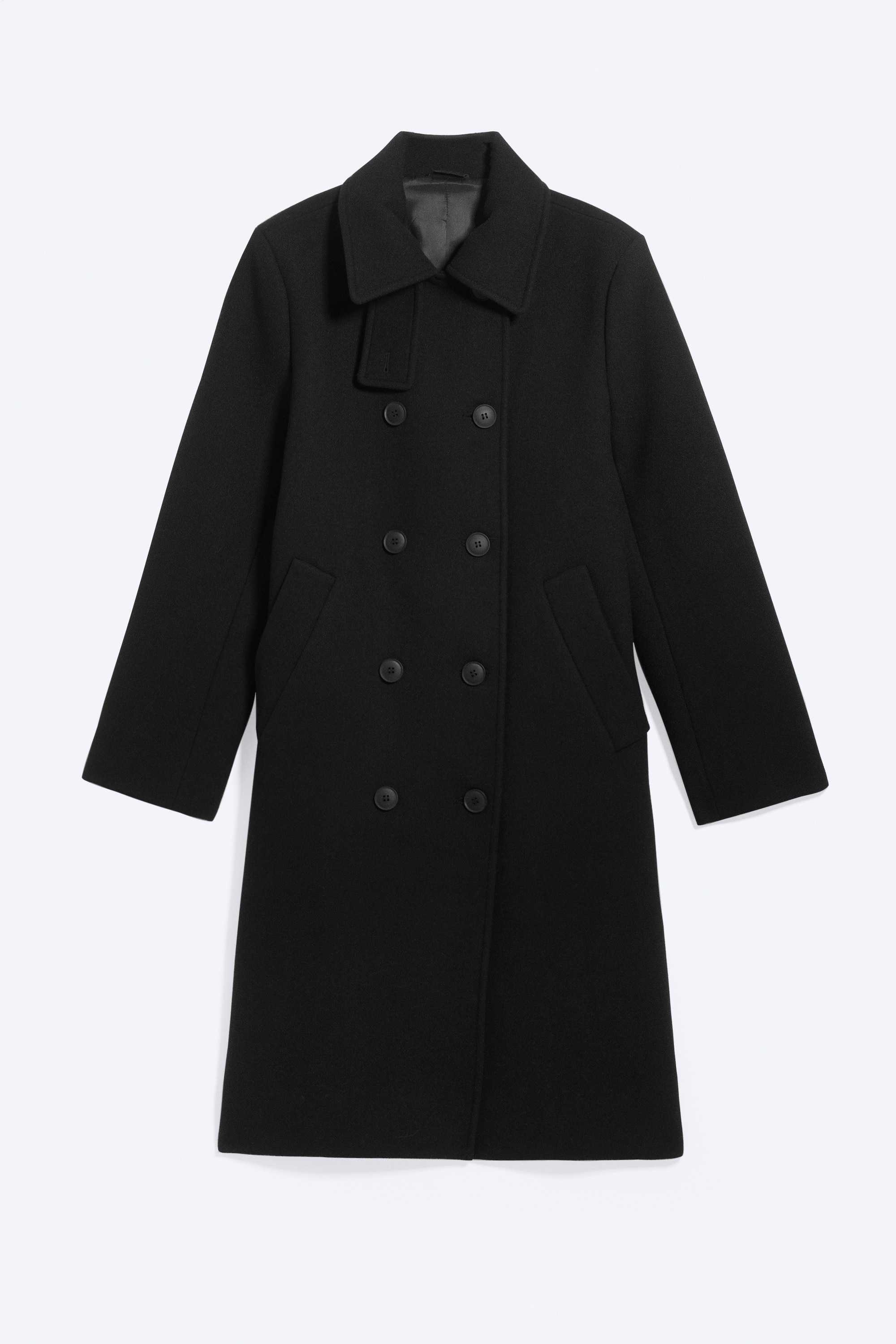 Modellen bÃ¤r en svart car coat i knÃ¤lÃ¤ngd med dubbelknÃ¤ppning och stÃ¥r med hÃ¤nderna instoppade i de snedstÃ¤llda passpoalfickorna. Den strukturerade kappan har en hÃ¶g, knÃ¤ppt krage och Ã¤r stylad med grÃ¥ jeans och svarta skor med spetsig tÃ¥.