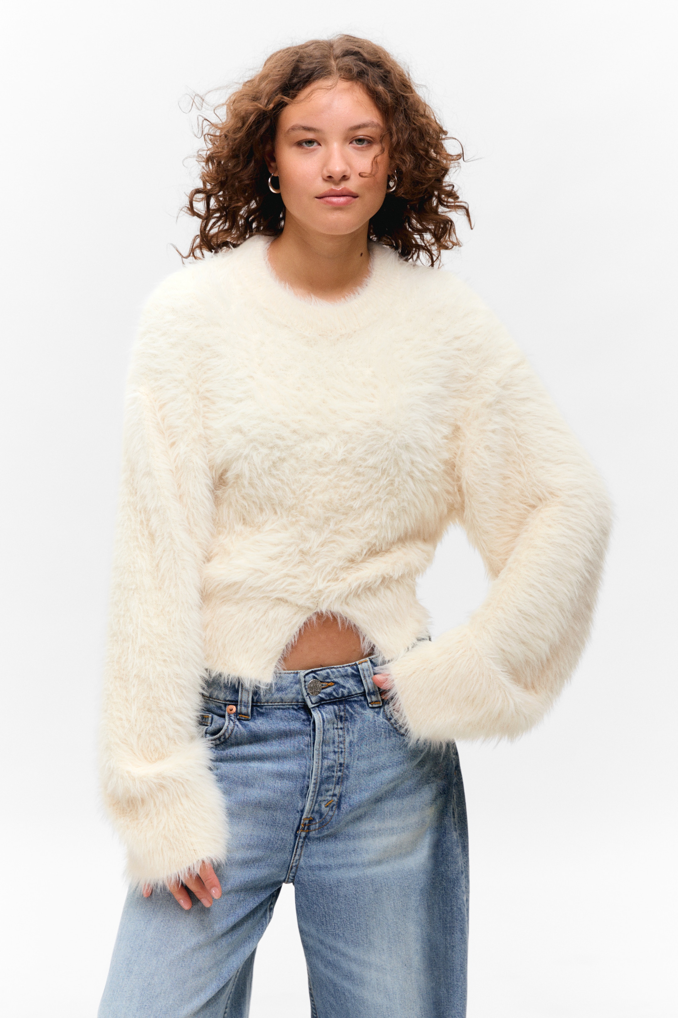 Ingrandisci l'immagine: Un maglione soffice color bianco sporco presenta un girocollo e maniche lunghe e ampie. Il design dell'orlo incrociato del maglione rivela una parte del ventre ed è abbinato a jeans lavaggio chiaro.