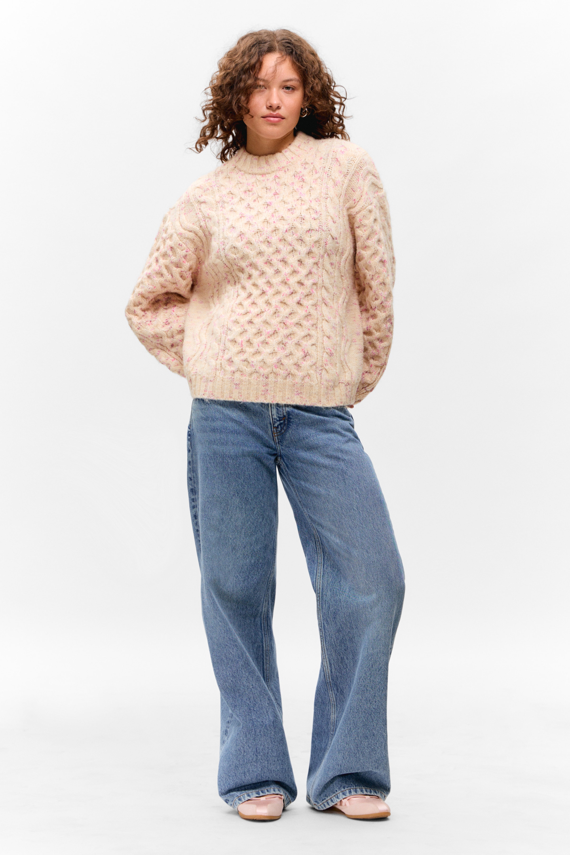 Visa större bild: En ljus krämfärgad kabelstickad tröja med rosa stänk presenteras, med en rund hals, ribbade muddar och ett texturerat honungskaka- och flätmönster. Den kombineras med ljusblå jeans med vida ben och glänsande rosa loafers.