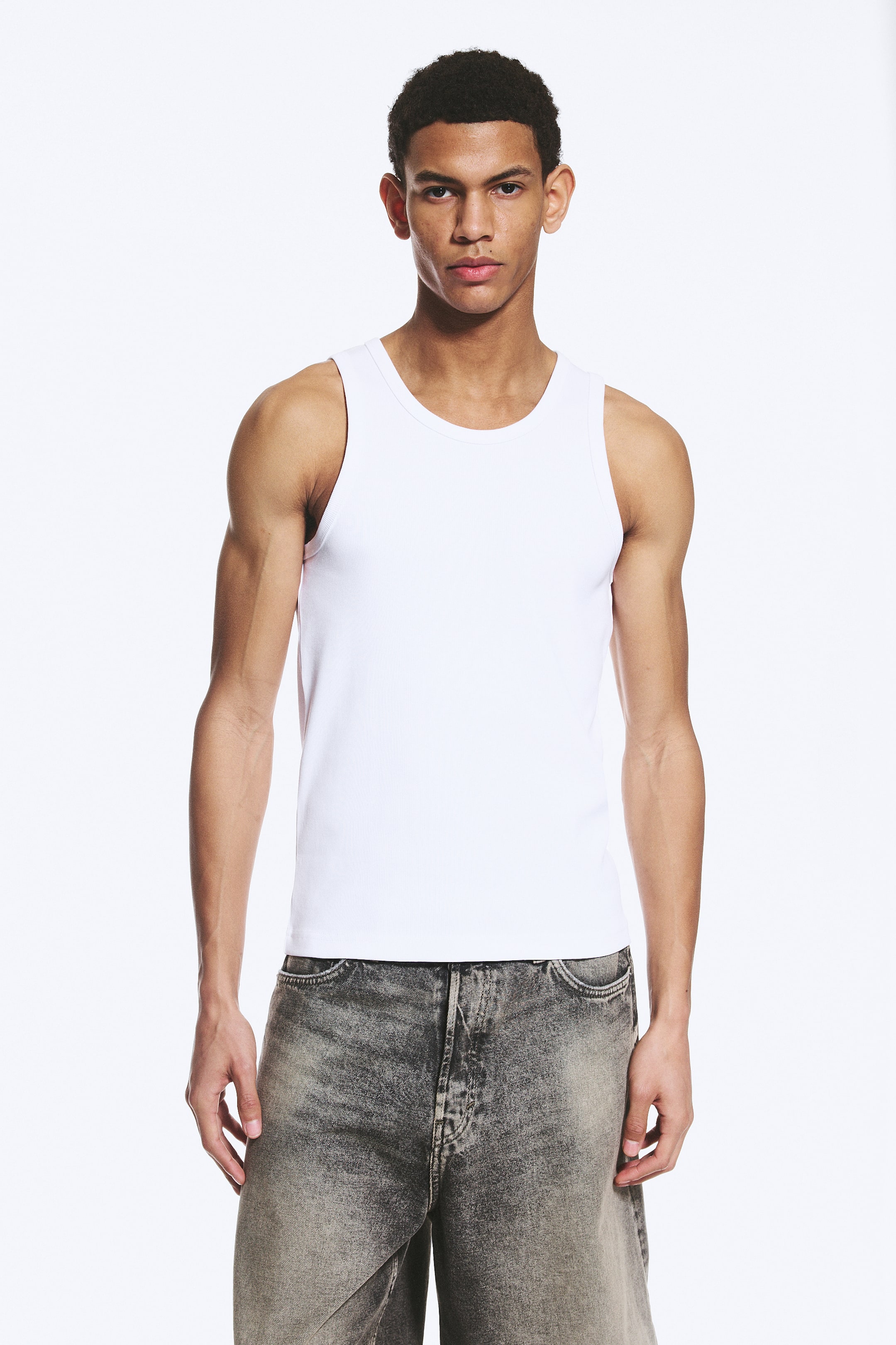 Grösseres Bild ansehen: Ein weißes Tanktop mit rundem Ausschnitt, breiten Armausschnitten und einer dezenten Rippstruktur. Es präsentiert sich in einem figurbetonten Design mit geradem Saum, frontseitig dargestellt.