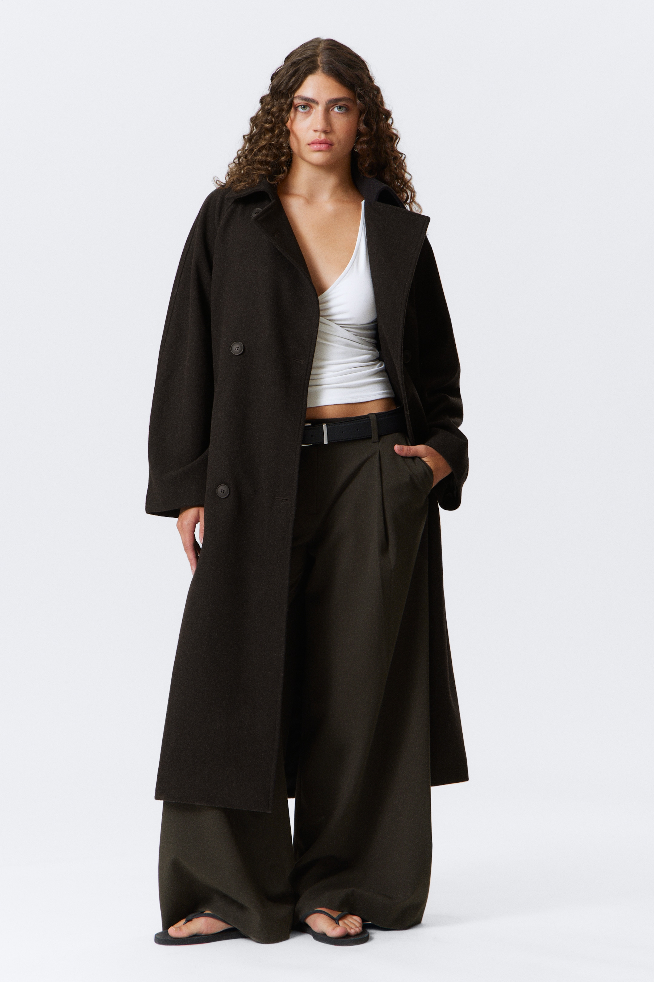 Agrandir l'image: Un manteau long de couleur brun foncé est présenté de face. Il est doté de larges revers crantés et d'un design croisé avec deux boutons visibles, offrant une coupe ample et décontractée. Le manteau est ouvert, révélant un haut blanc et un pantalon large de couleur sombre en dessous.
