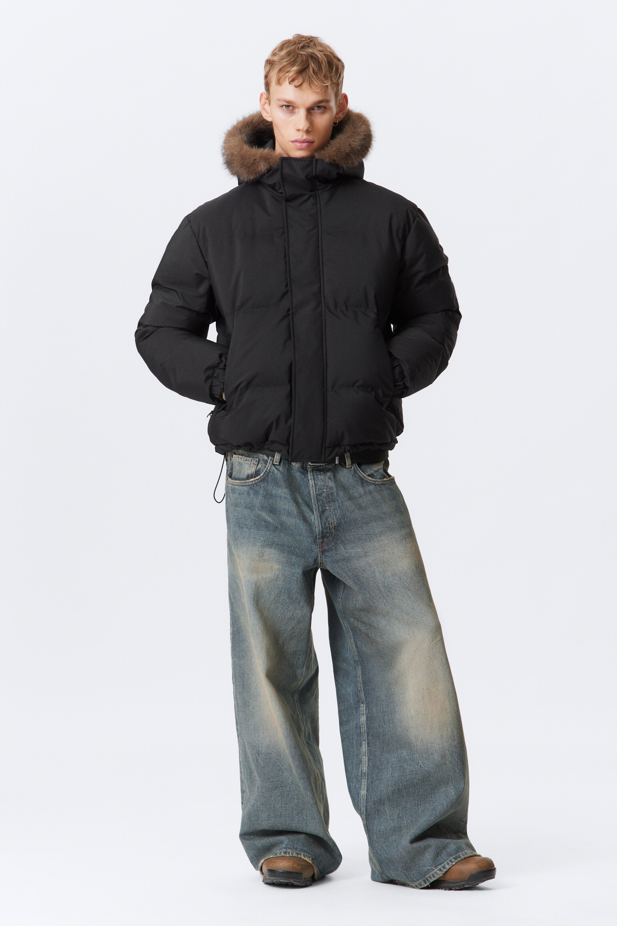 Grotere afbeelding bekijken: Een vooraanzicht van een zwarte gewatteerde jas met een kap met bruine bontafwerking, een gewatteerd ontwerp en een voorritssluiting. Het wordt gedragen over wijde, vaalblauwe jeans.