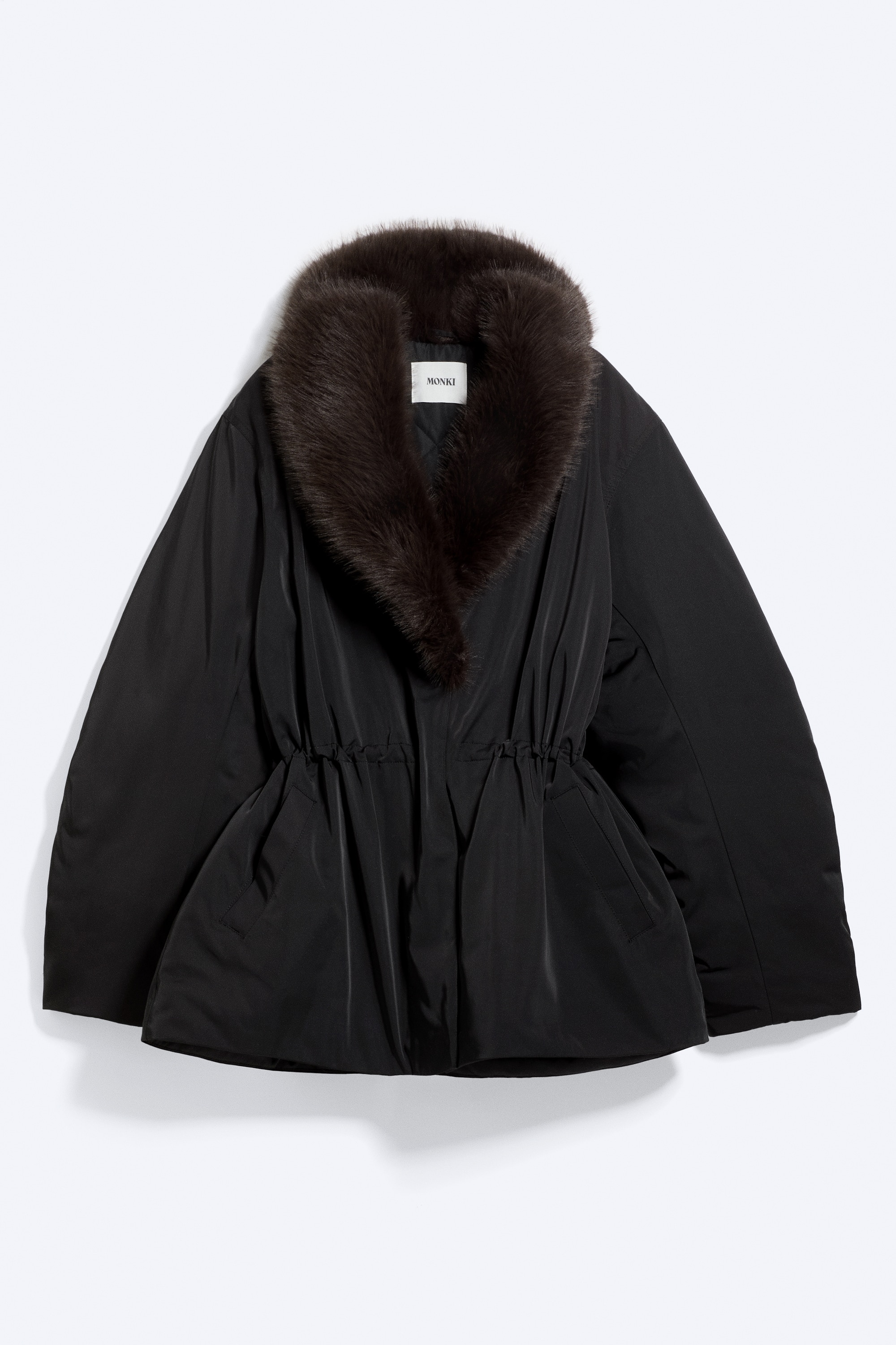 A black padded jacket featuring a dark faux fur shawl collar and a cinched elasticated waist is being worn by the model. The jacket has a concealed front fastening and voluminous sleeves, styled with matching black trousers.