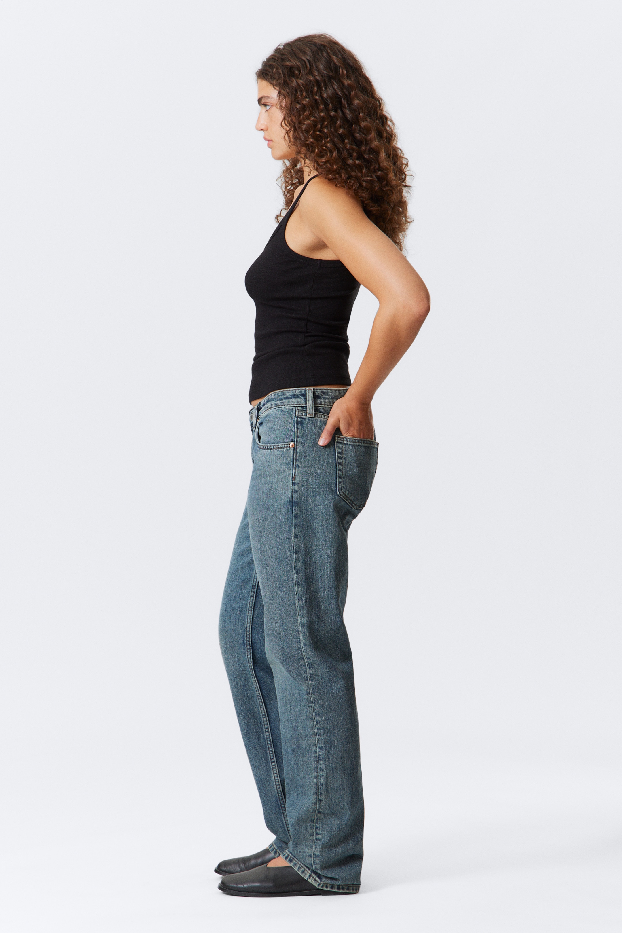Grösseres Bild ansehen: Ein schwarzes geripptes Tanktop wird getragen, das dünne Träger und eine figurbetonte Silhouette aufweist. Es wird mit mittelblauen Jeans im Used-Look gestylt, die eine entspannte, gerade Passform aus der Seitenansicht zeigen, einschließlich sichtbarer Fronttaschen und Gürtelschlaufen.