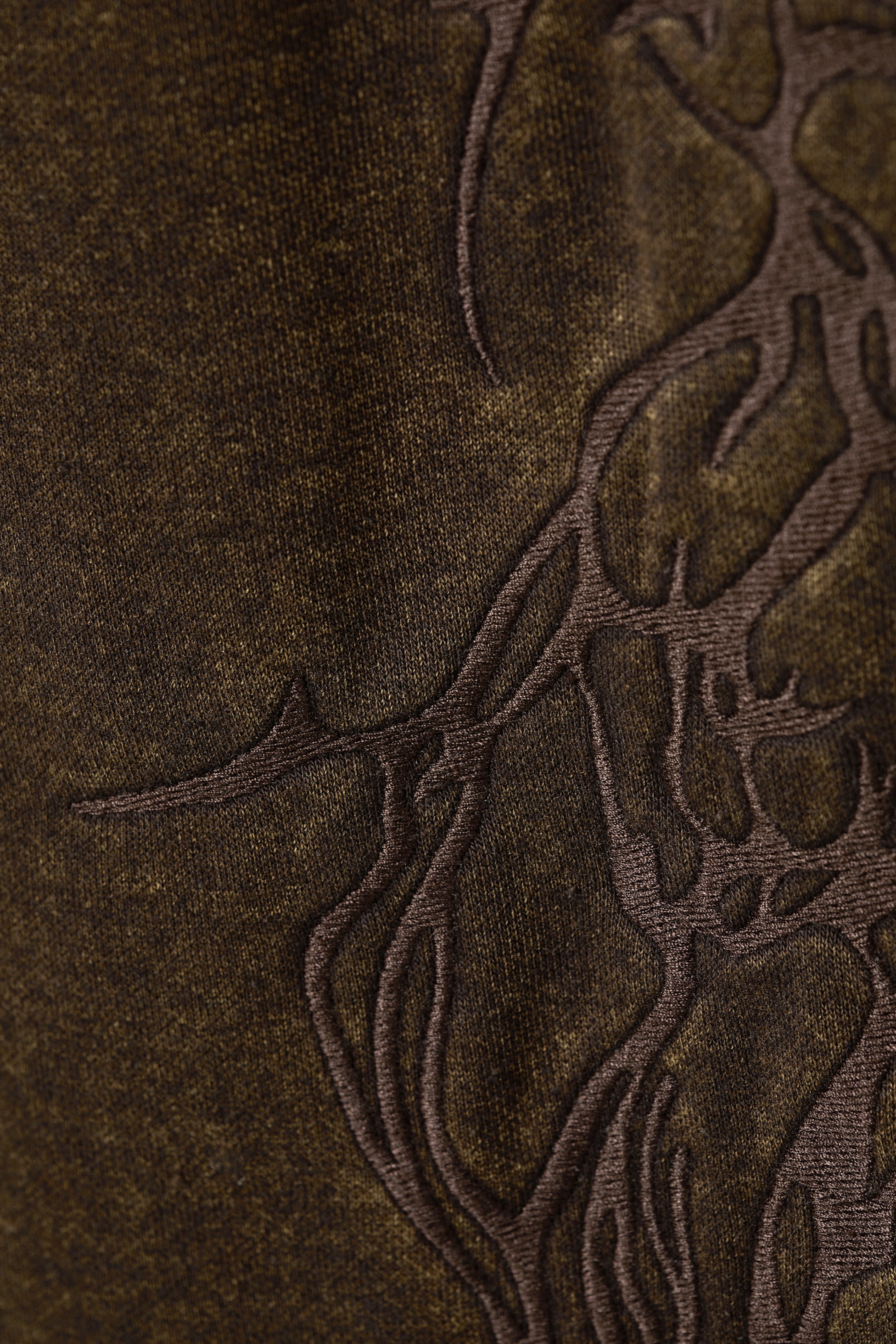 Ver imagem maior: Uma calça de moletom de algodão em verde oliva escuro está a mostrar um close-up da textura manchada do seu tecido. Um bordado intricado em castanho escuro está a formar um padrão orgânico, semelhante a raízes, ao longo do tecido.
