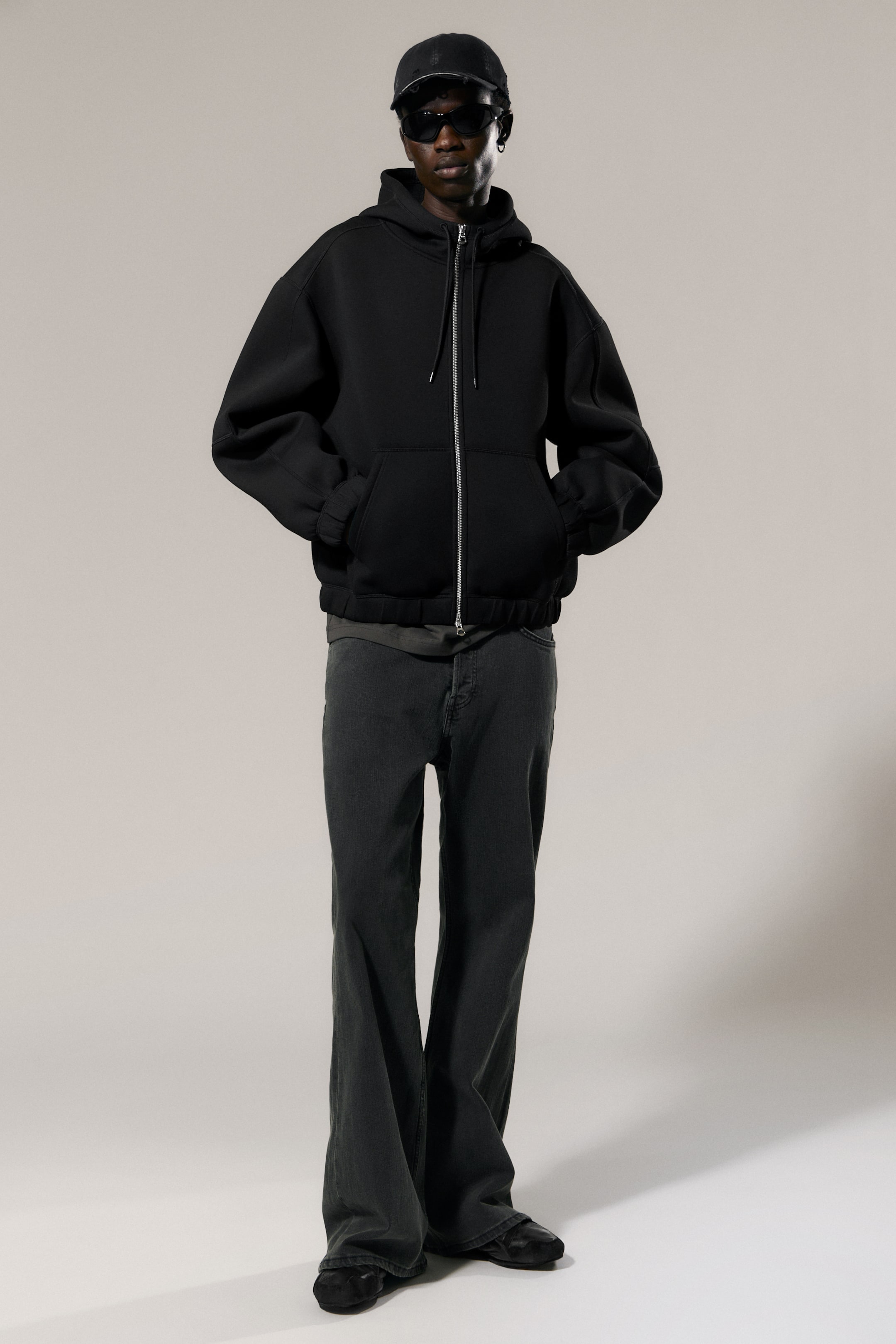 View larger image: A black full-zip hoodie presents an oversized fit and features a structured hood, a silver-toned front zip, and large front pockets where hands can rest. It also displays elasticated cuffs and hem, with a grey T-shirt layered underneath.