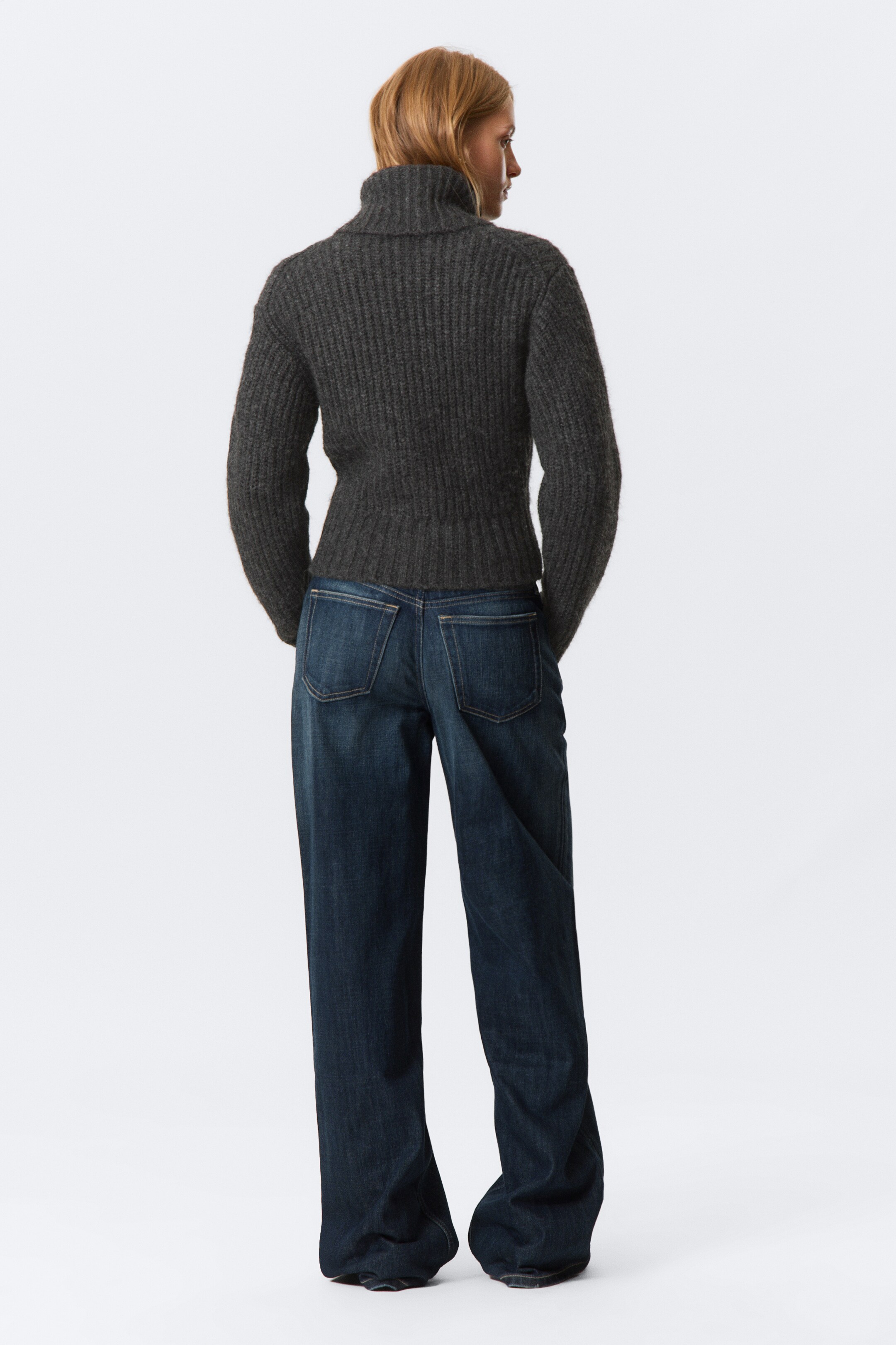 Visa större bild: En mörkgrå stickad kofta med ribbad textur, bred vikt krage, åtsittande ärmar och tajta ribbade kanter bärs av modellen. Modellen står med händerna bakom ryggen och stylar koftan med vida jeans.
