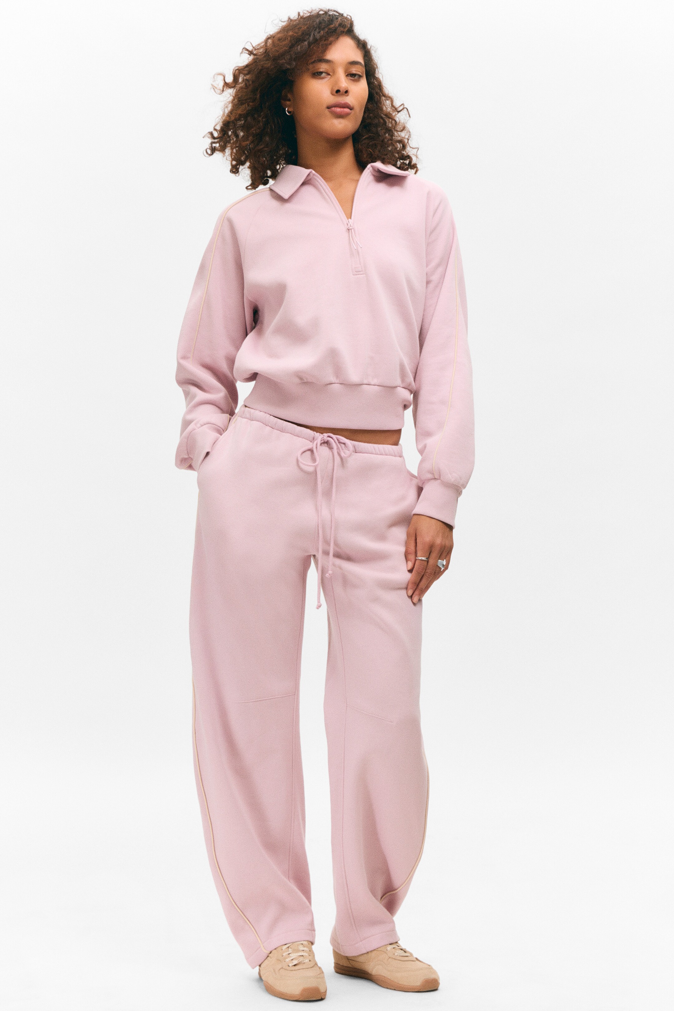 Agrandir l'image: Un survêtement rose est présenté, comprenant un haut court à demi-zip et un pantalon à jambes larges, tous deux avec un détail de passepoil de couleur claire le long des coutures extérieures. Le haut est conçu avec un col pointu, une fermeture éclair argentée à demi-zip, et des poignets et une base côtelés, tandis que le pantalon assorti est doté d'une taille élastique avec cordon de serrage.