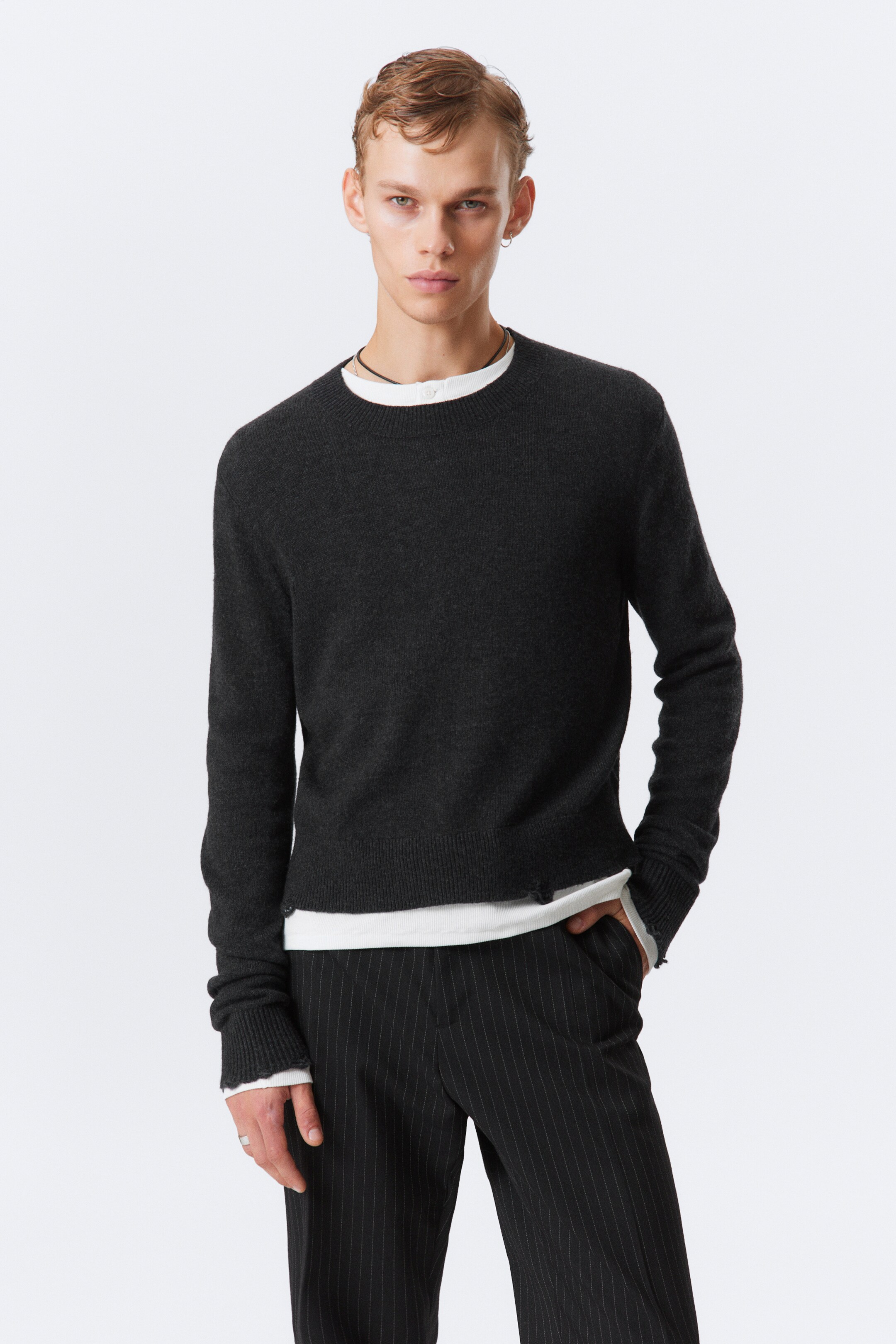 View larger image: A dark grey crew neck sweater is being presented, facing forward. It is featuring a fine knit texture, long sleeves with ribbed cuffs, and a rolled bottom hem, revealing a white undershirt peeking out.