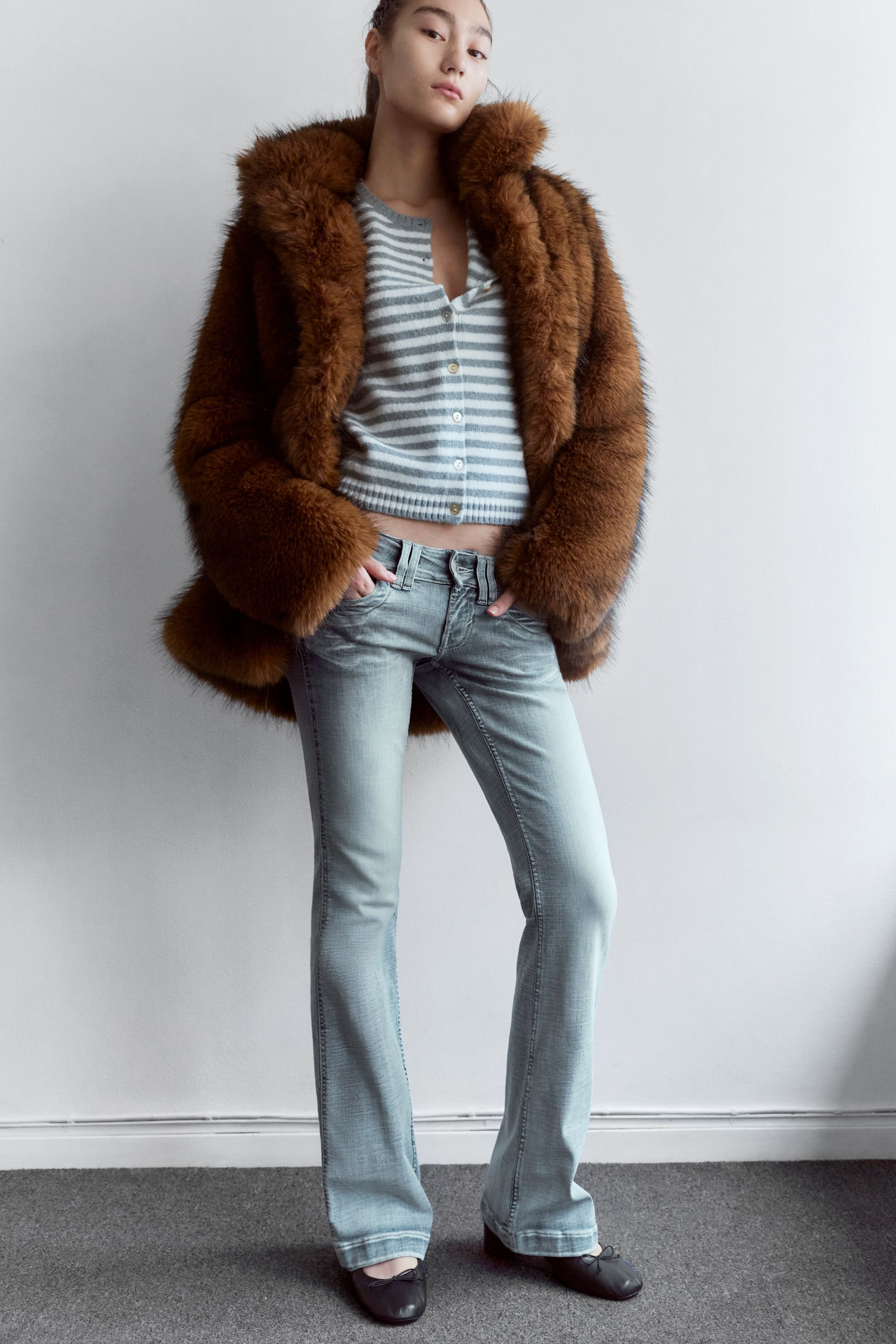 View larger image: Light blue, low-rise jeans with a slim fit and flared leg are being worn by the model, who is standing with hands resting in the front pockets. The jeans are styled with a striped cardigan, a brown faux fur coat, and black ballet pumps.