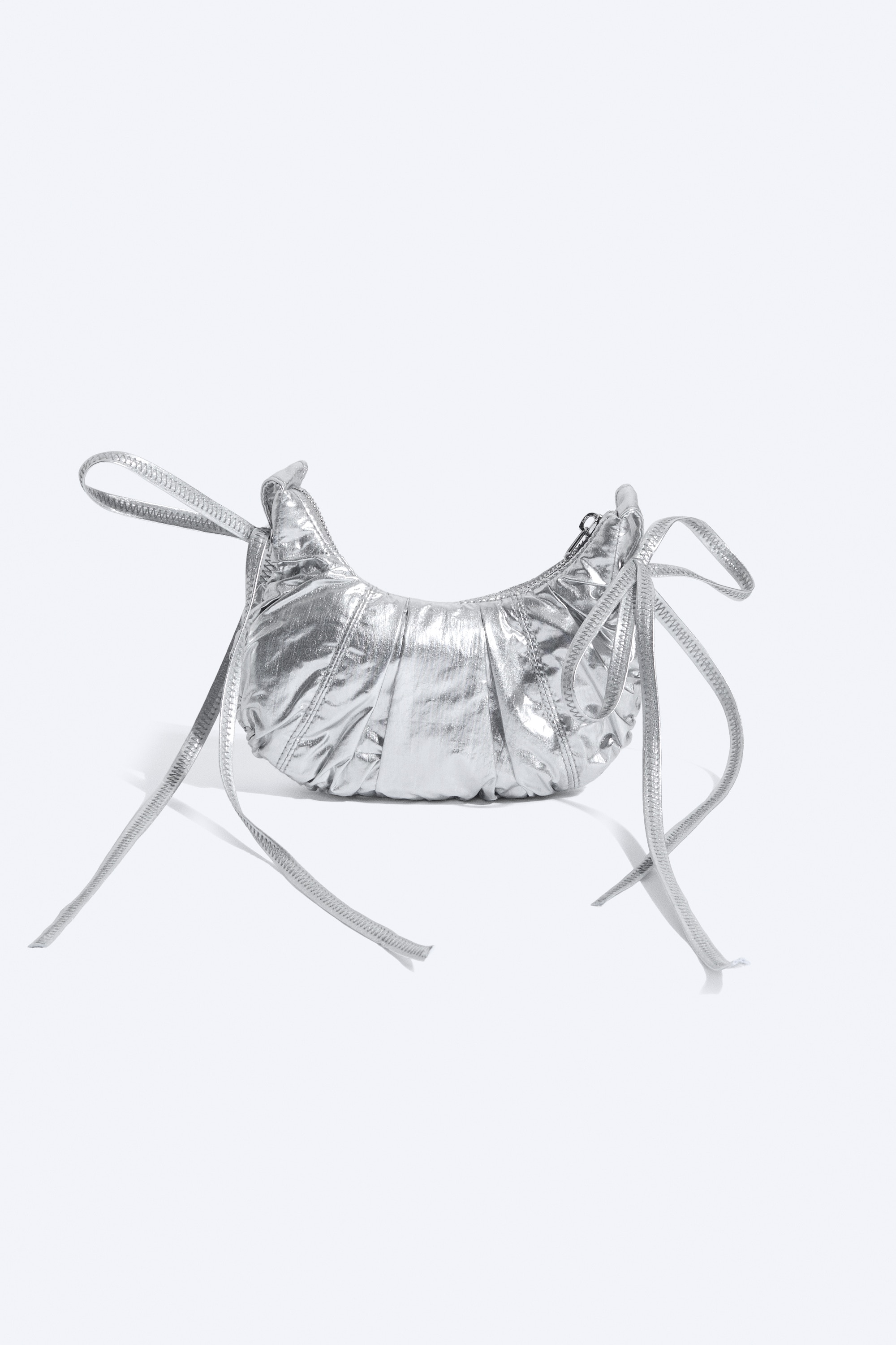Malá zakřivená crossbody kabelka v metalické stříbrné barvě s řasenou texturou a dlouhými splývavými stuhami je nošena přes rameno modelky. Kabelka spočívá na krémovém pleteném svetru, čímž vyniká její lesklý, odrazivý povrch.
