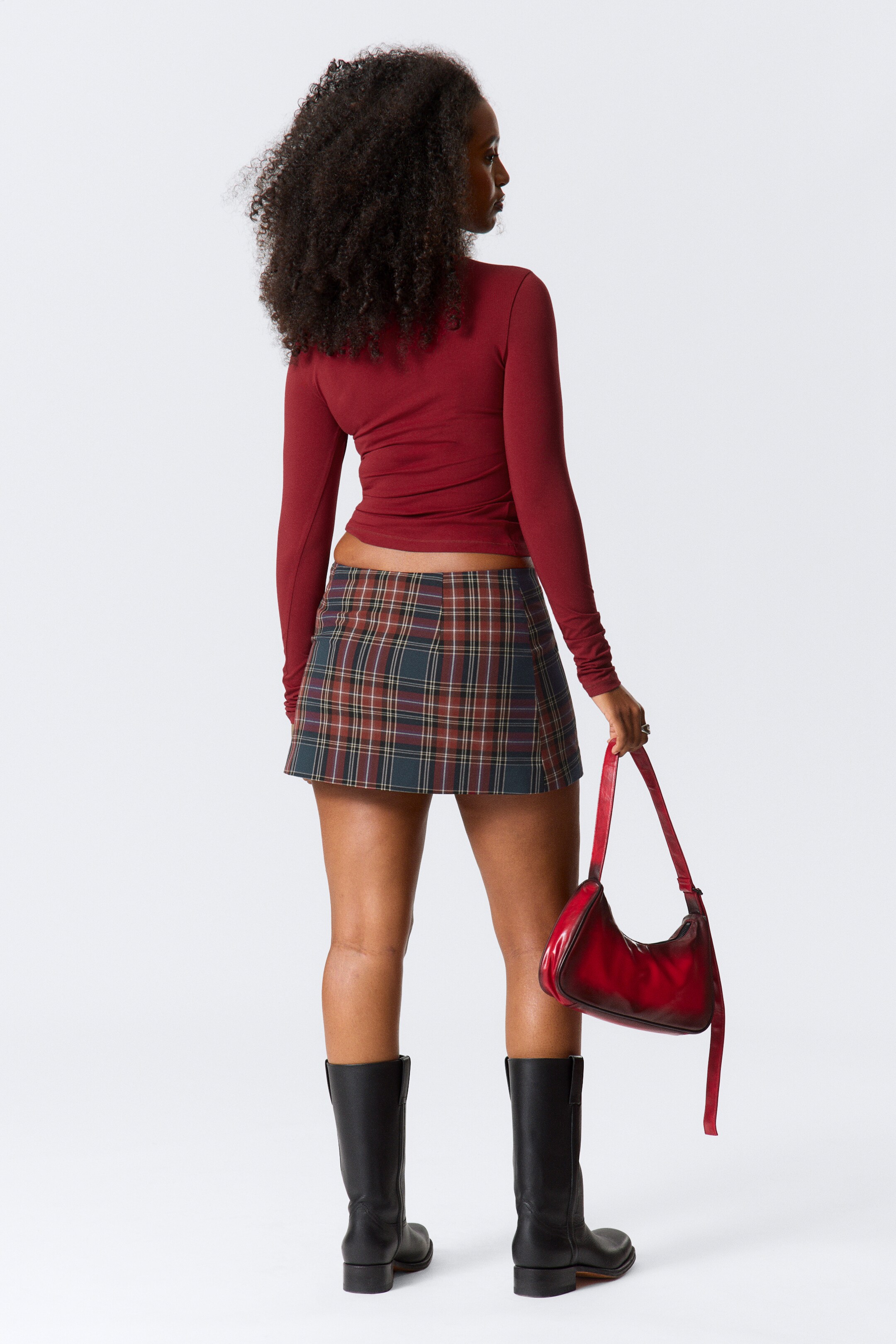 View larger image: A dark red, long-sleeved, form-fitting top is being worn, cropped at the waist and viewed from the back, paired with a plaid mini skirt featuring red, green, and brown hues. A red, distressed-look crescent shoulder bag is being held, while black mid-calf boots are completing the outfit.
