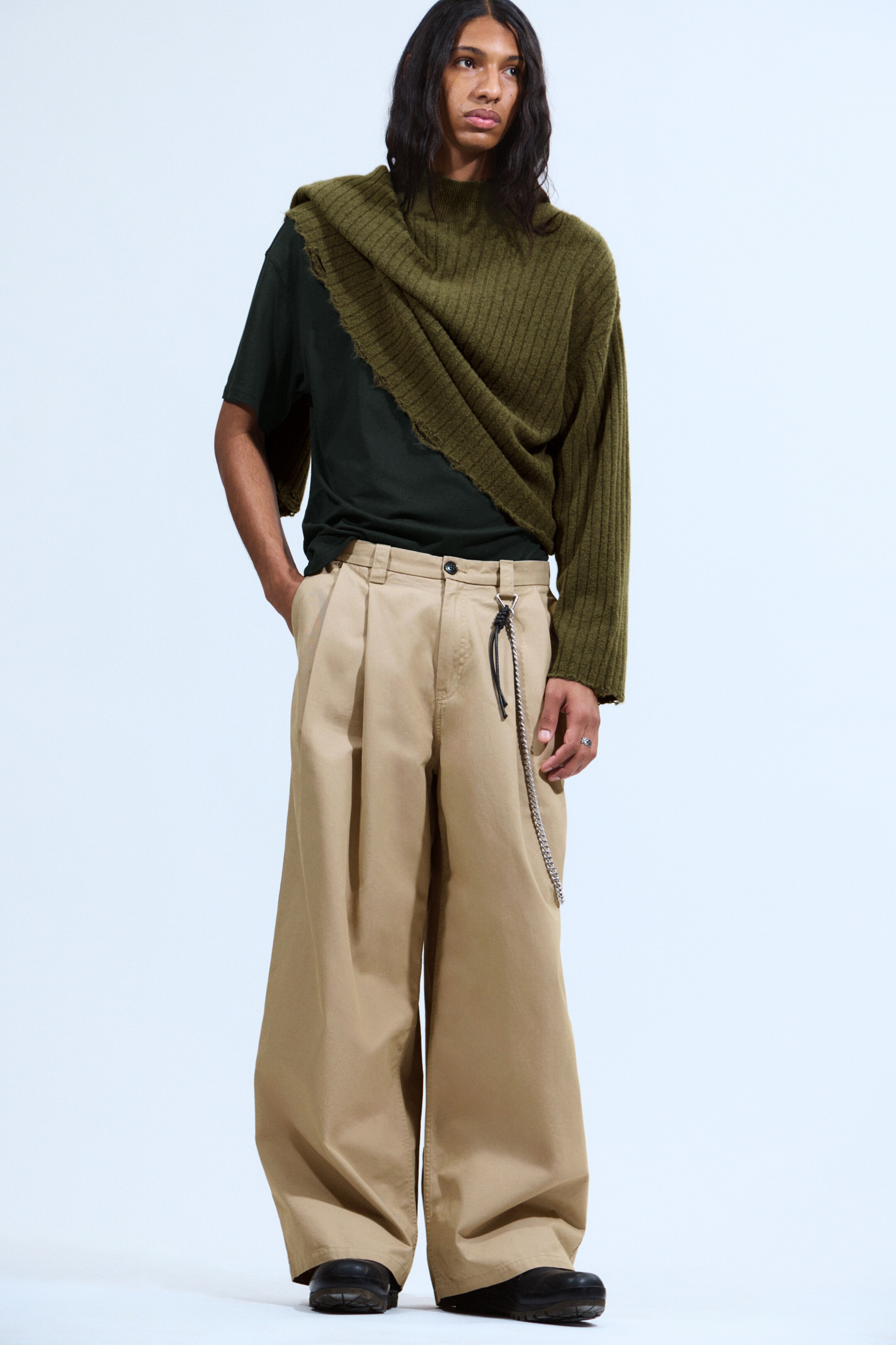 Se større billede: Et komplet outfit præsenteres, med en olivengrøn, ribstrikket sweater med høj hals, en lang ærme og en asymmetrisk råkantet søm, lagdelt over en mørkegrøn kortærmet t-shirt. Brede, plisserede khakibukser bæres, fremhævet af en sølvkæde, og fuldendt med sorte chunky sko.