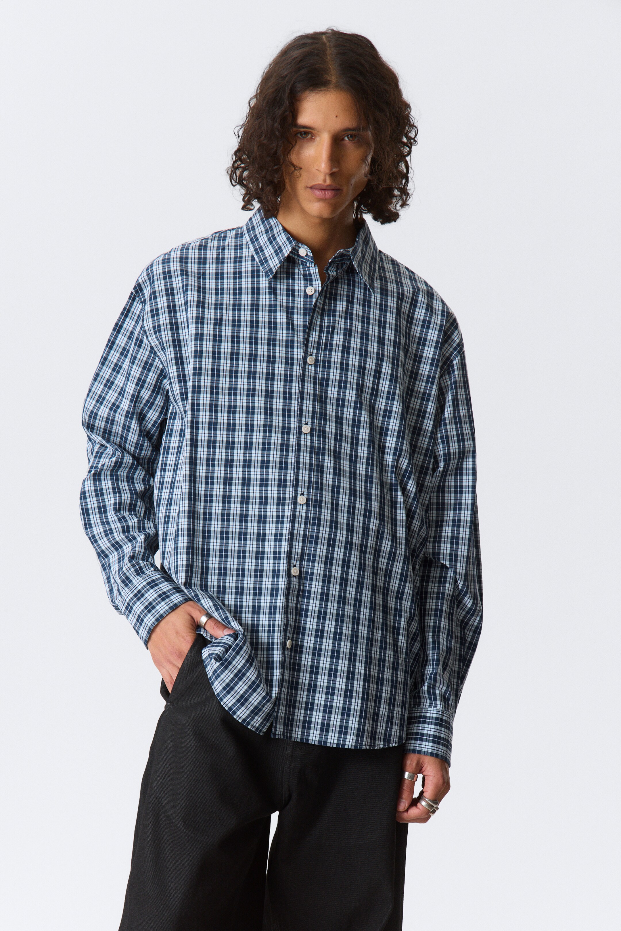 View larger image: A long-sleeved shirt is displaying a blue, dark blue, and white plaid pattern. It is designed with a pointed collar, a button-front closure featuring white buttons, and long sleeves ending in buttoned cuffs, presenting an oversized fit.