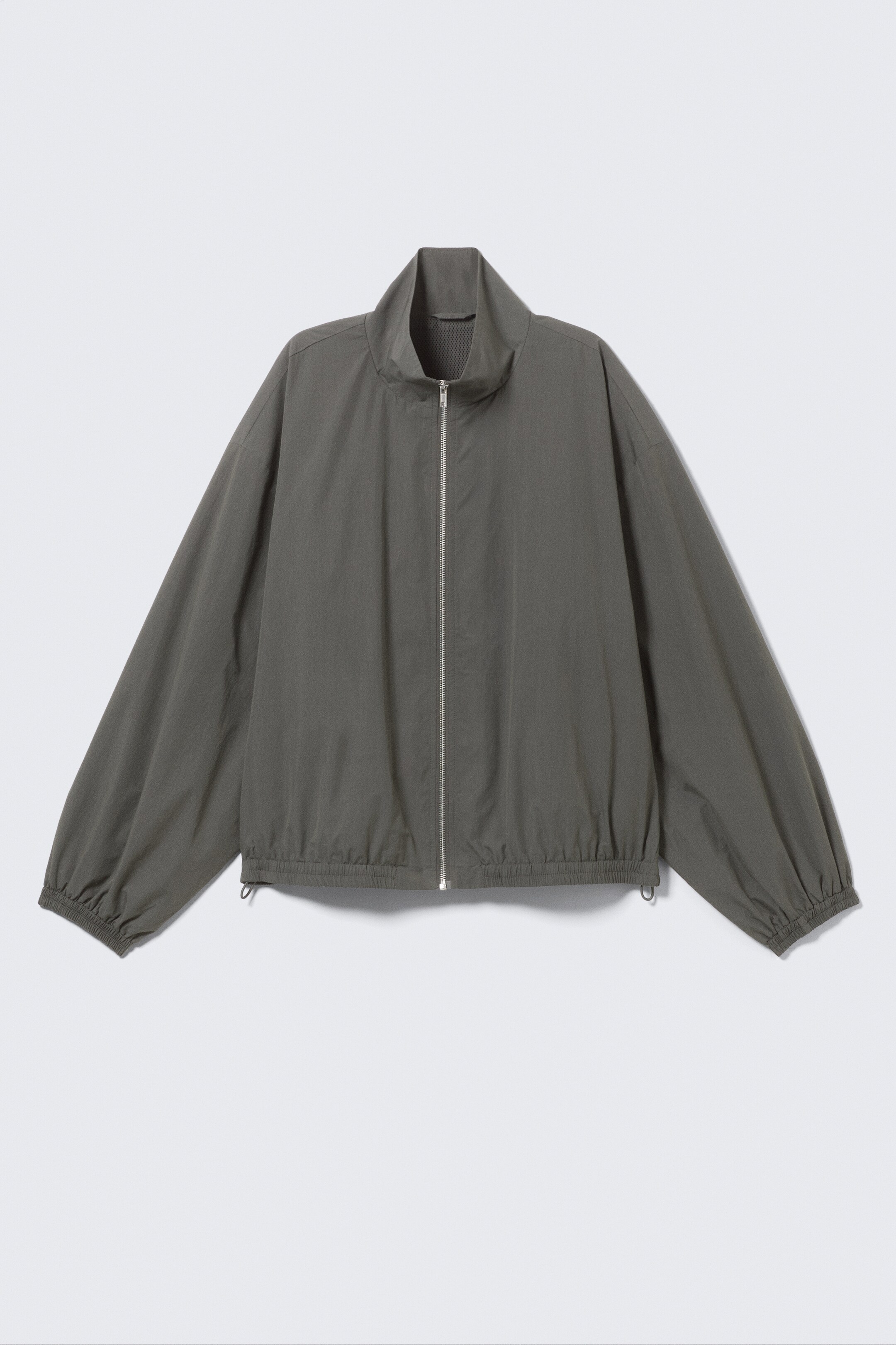 Преглед на по-голямо изображение: Овърсайз трак яке в каки-зелен цвят е показано отпред, с висока яка, цип по цялата дължина със сребрист оттенък, еластични маншети и еластичен долен подгъв с видими връзки.