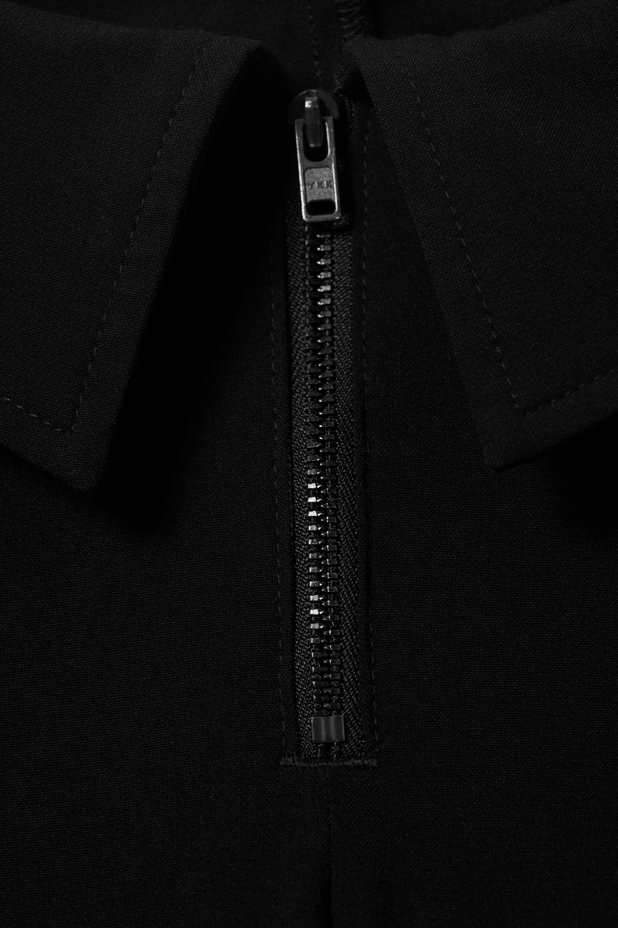 Agrandir l'image: Un vêtement noir est doté d'un col pointu et d'une fermeture éclair YKK de couleur argent partiellement ouverte qui descend le DEVANT.