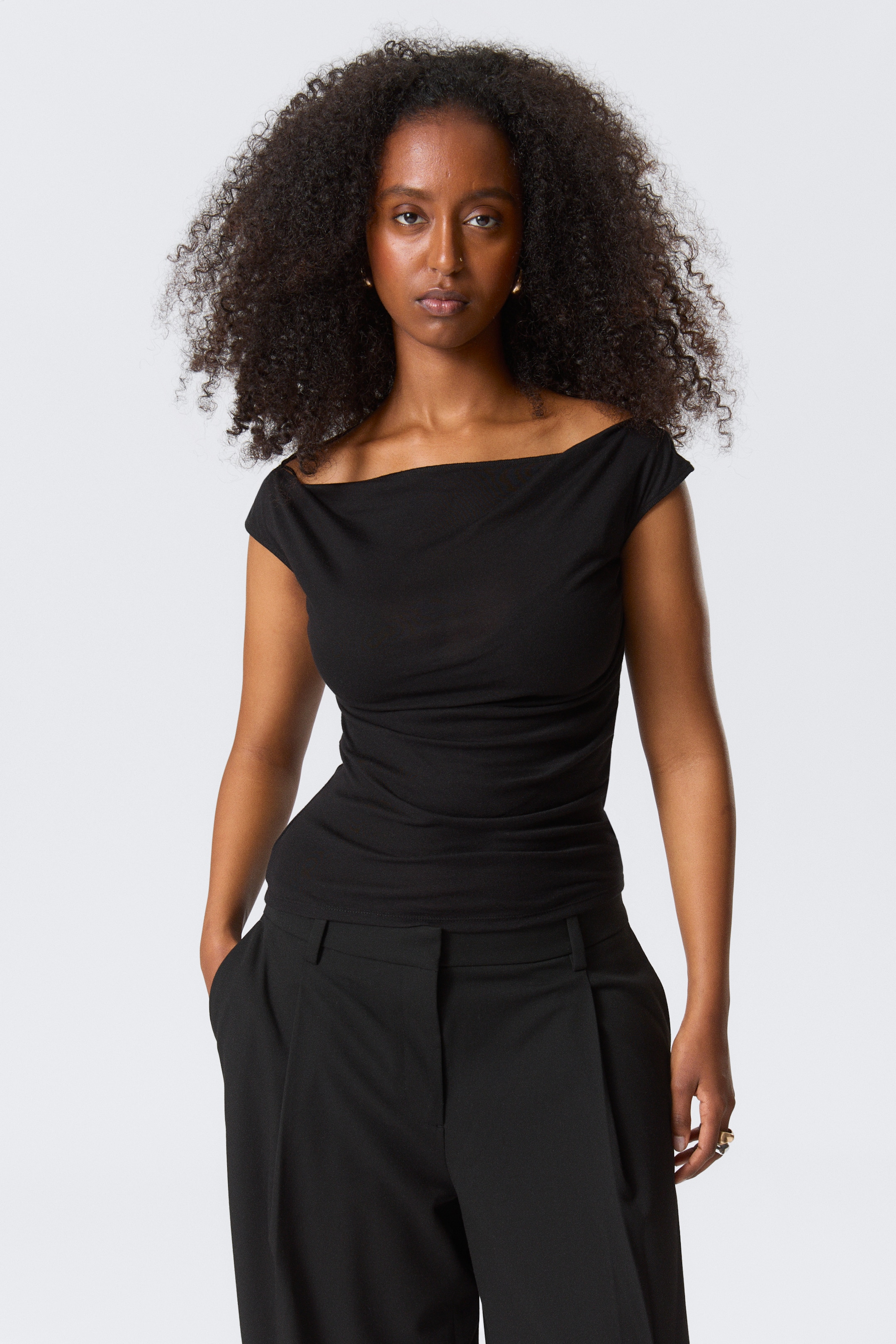Agrandir l'image: Un haut noir sans manches, avec une silhouette à larges épaules et un col drapé, est porté par le mannequin. Le haut ajusté présente des plis subtils le long d'un côté de la taille, et le mannequin se tient avec une main dans la poche d'un pantalon noir.