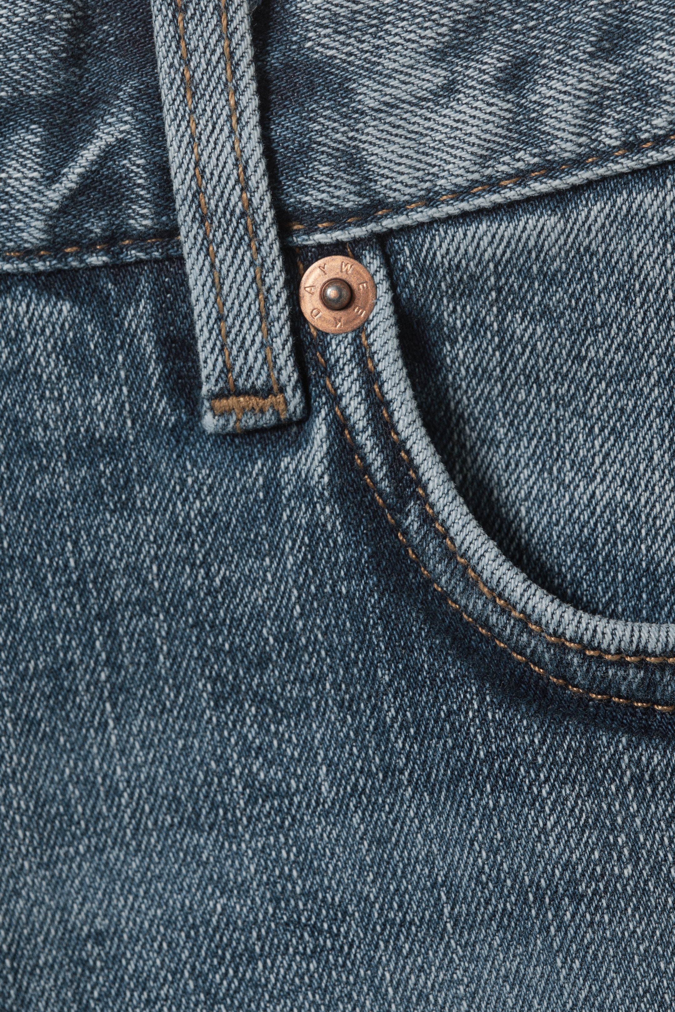 Agrandir l'image: Un jean en denim bleu est présenté en gros plan sur le côté supérieur avant droit. Un rivet en métal de ton cuivre sécurise le coin d'une poche avant incurvée, qui est détaillée avec une double couture contrastante orange. Un passant de ceinture est également visible à côté du rivet.