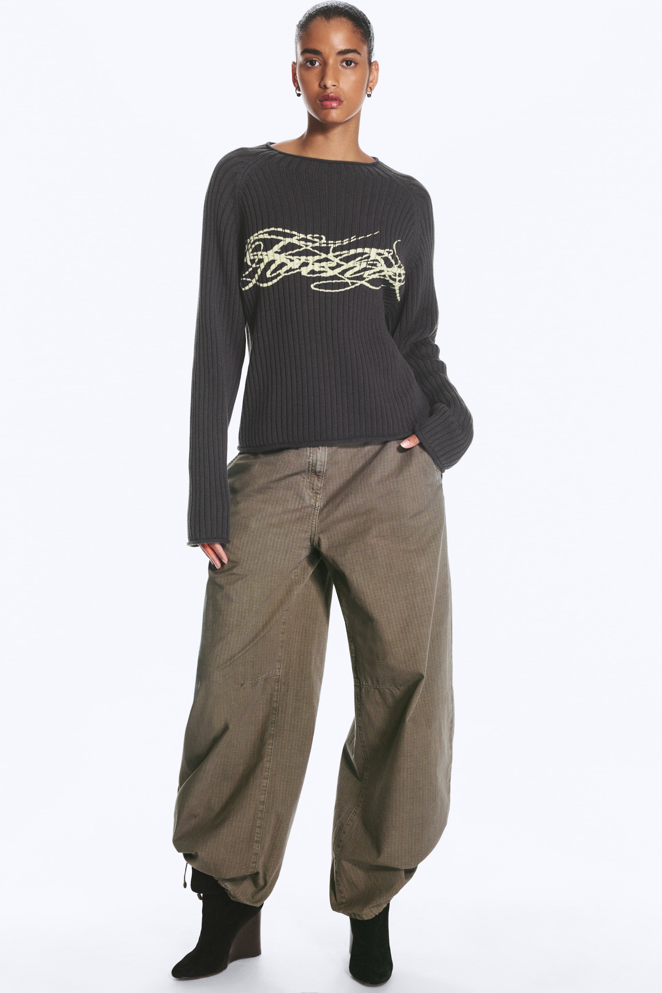 Agrandir l'image: Un pull en maille côtelée épaisse gris foncé, doté d’un col montant imitation, de manches raglan et d’un imprimé graphique jaune clair sur la poitrine, est porté par le mannequin. Le pull présente des bords roulés à l’encolure, aux poignets et à l’ourlet, et est associé à un pantalon large.