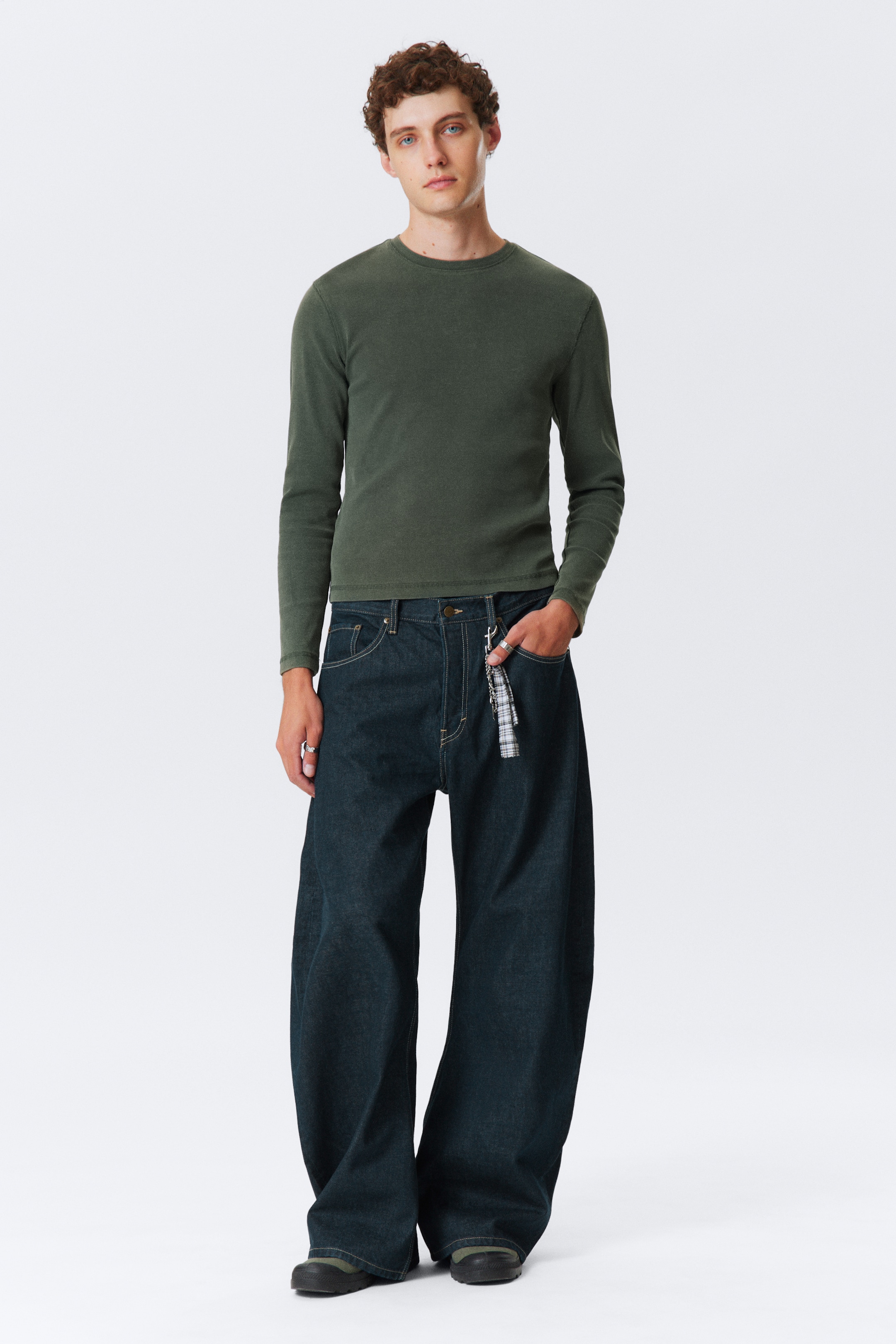 Visa större bild: En mörk olivgrön långärmad tröja med rund hals sitter tätt på överkroppen och armarna. Den kombineras med mörktvättade, vida jeans som har en lös, avslappnad passform och samlas vid anklarna.