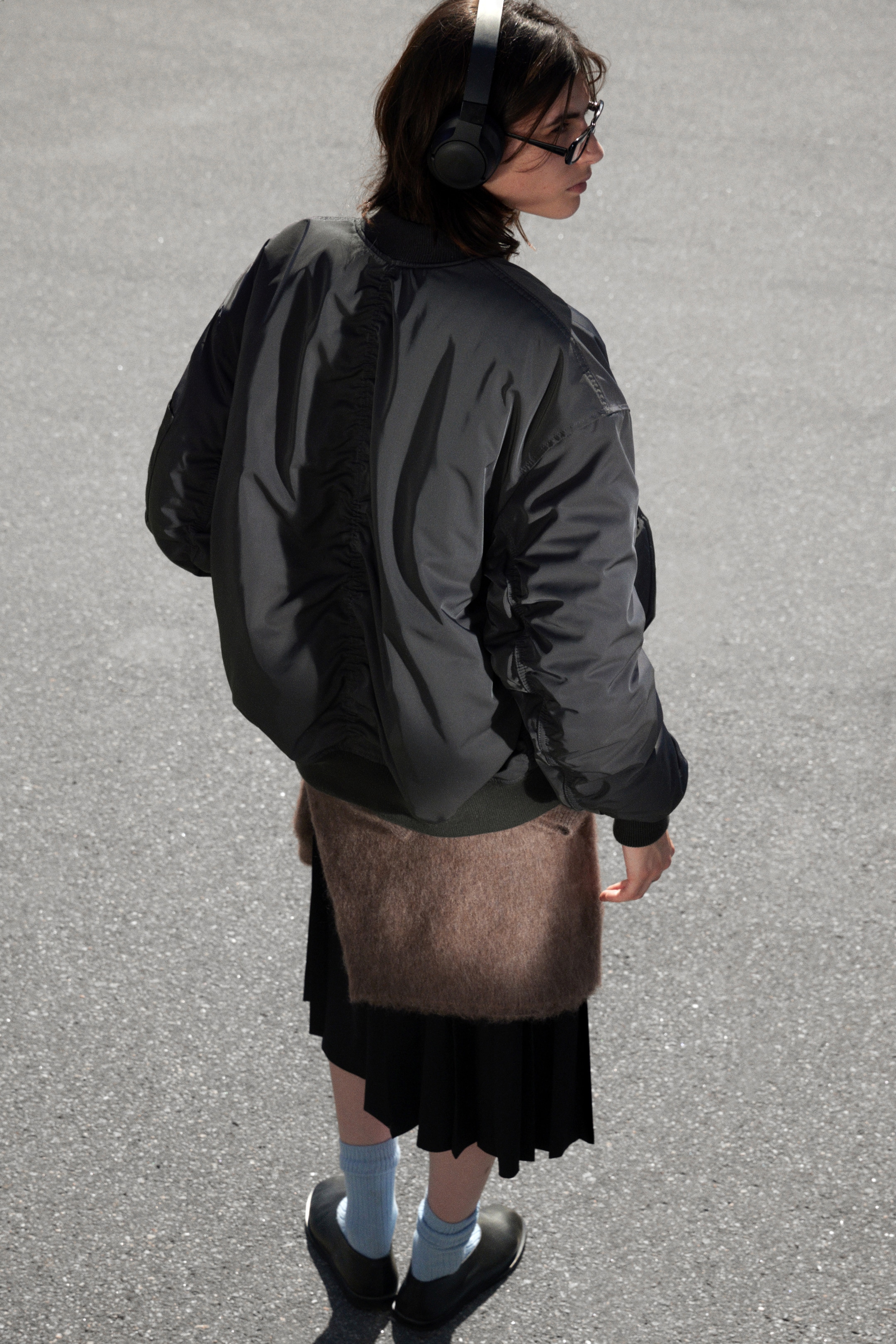 View larger image: A dark grey bomber jacket is being viewed from the back-right angle, featuring a ribbed collar, cuffs, and hem. Its full, slightly puffed sleeves and back seams are showcasing gathering details, emphasizing the jacket's relaxed fit.