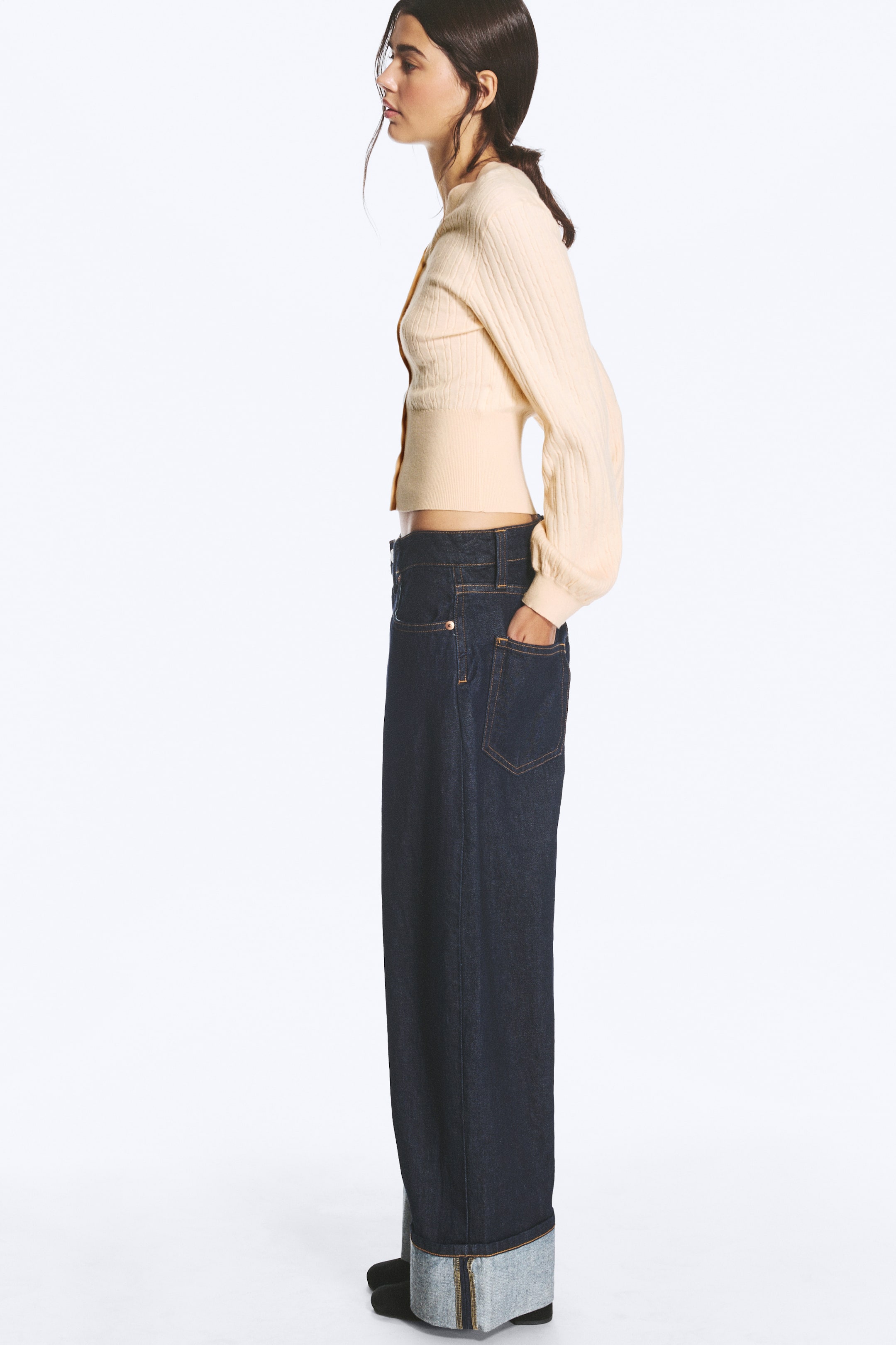 Visualizza l'immagine ingrandita: Un paio di jeans scuri, a vita bassa, con vestibilità ampia, gamba baggy e ampi risvolti è indossato dalla modella. La modella è in piedi di profilo, con una mano nella tasca posteriore, abbinata a un cardigan corto color crema che mette in risalto la vita bassa dei jeans.